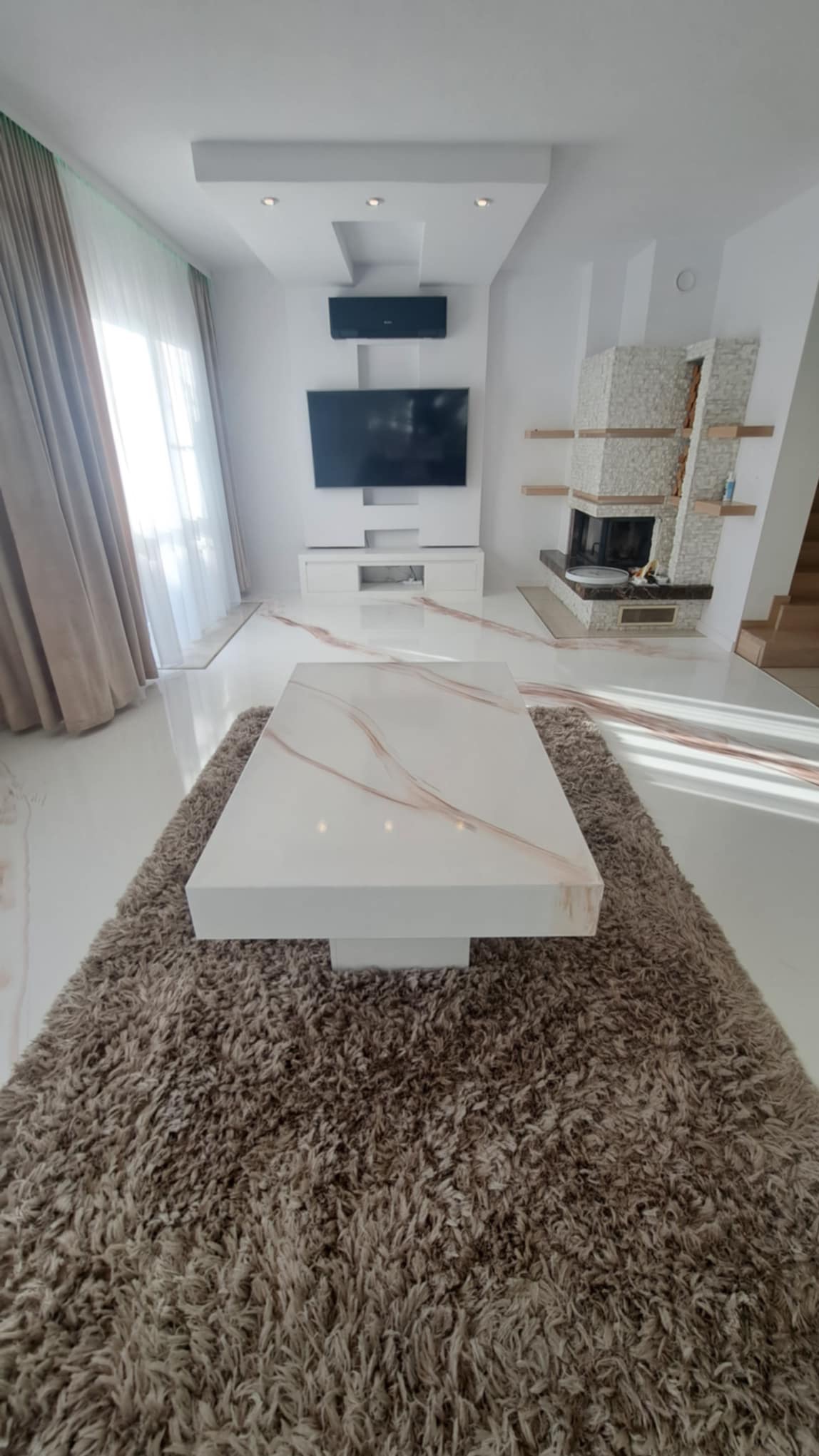 Jasne wnętrze salonu z marmurową podłogą i kominkiem obłożonym kamieniem, centralnie ustawiony biały stolik kawowy na puszystym, brązowym dywanie.