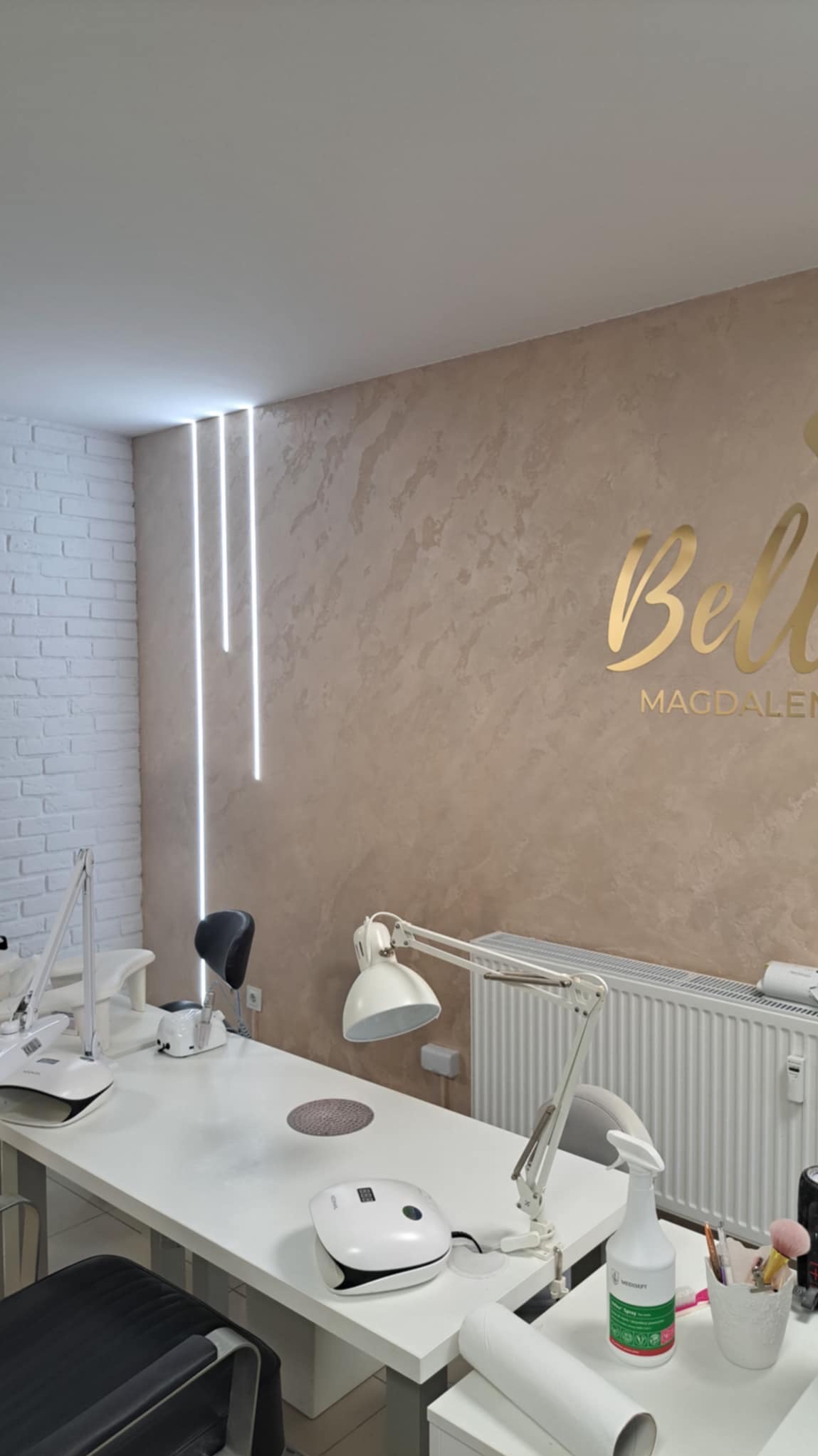 Stylowe wnętrze salonu kosmetycznego z białymi stolikami do manicure, lampami, urządzeniami UV i dekoracyjną ścianą z efektem trawertynu oraz podświetleniem LED.