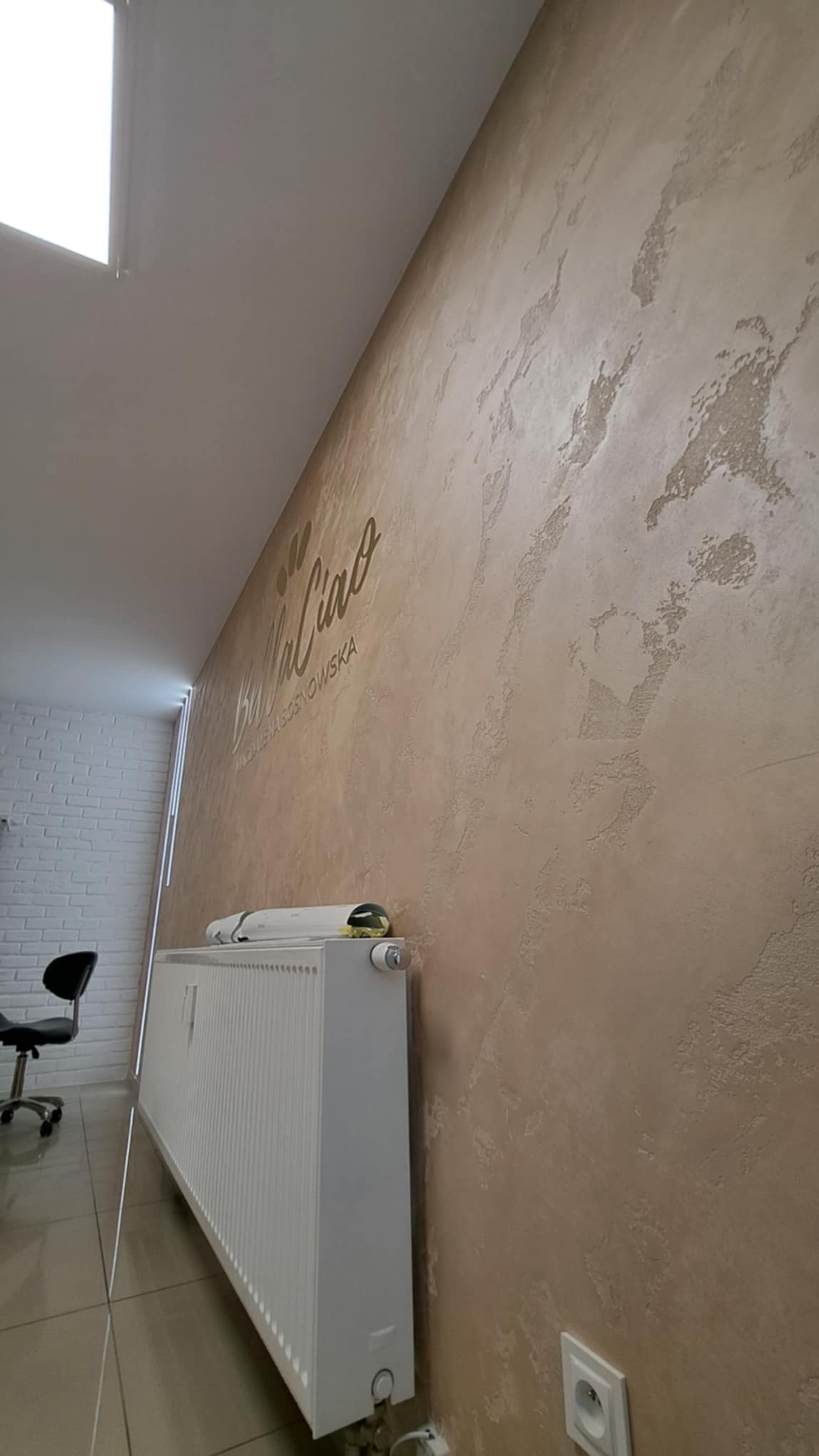 Dekoracyjna ściana z efektem stiuku w odcieniach beżu, z subtelnym napisem 'Bella Ciao', widoczny grzejnik i fragment biura z białymi cegłami w tle.
