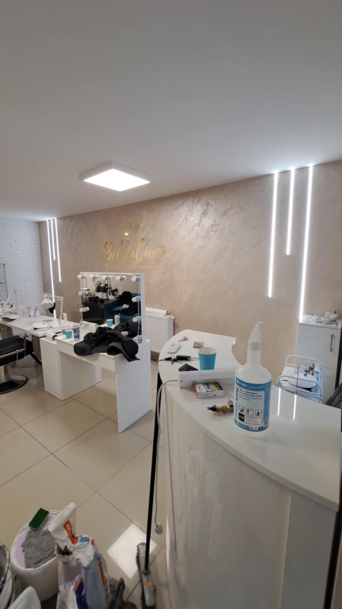 Wnętrze salonu kosmetycznego z białymi meblami, dekoracyjną ścianą z napisem 'Bella Ciao' i oświetleniem LED, widoczne akcesoria kosmetyczne i środki czystości.