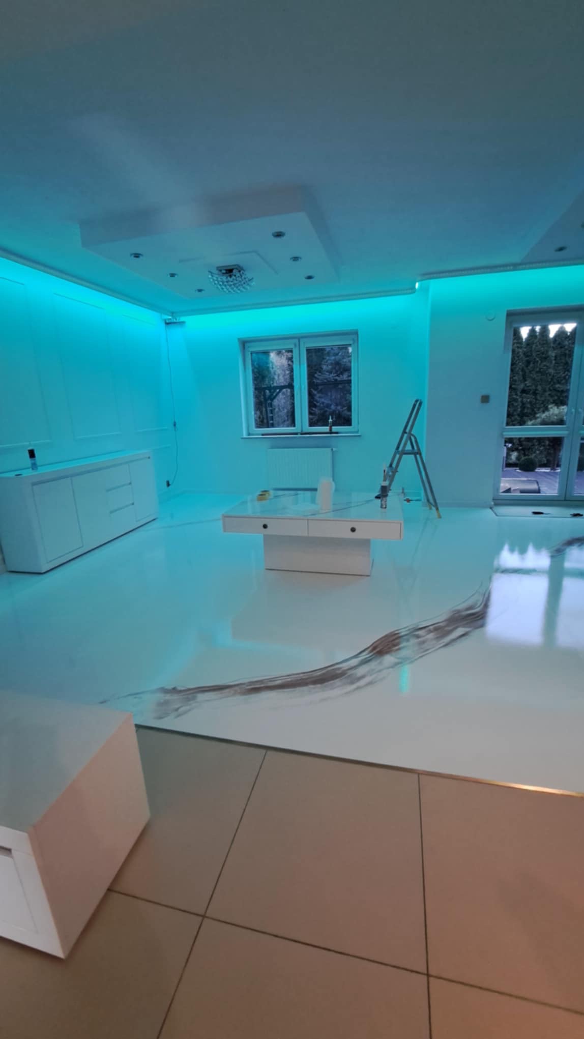 Białe wnętrze z żywiczną posadzką w stylu fali, oświetlone niebieskim światłem LED w Nowym Dworze Mazowieckim, z widoczną drabiną malarską i przygotowanymi powierzchniami ścian.