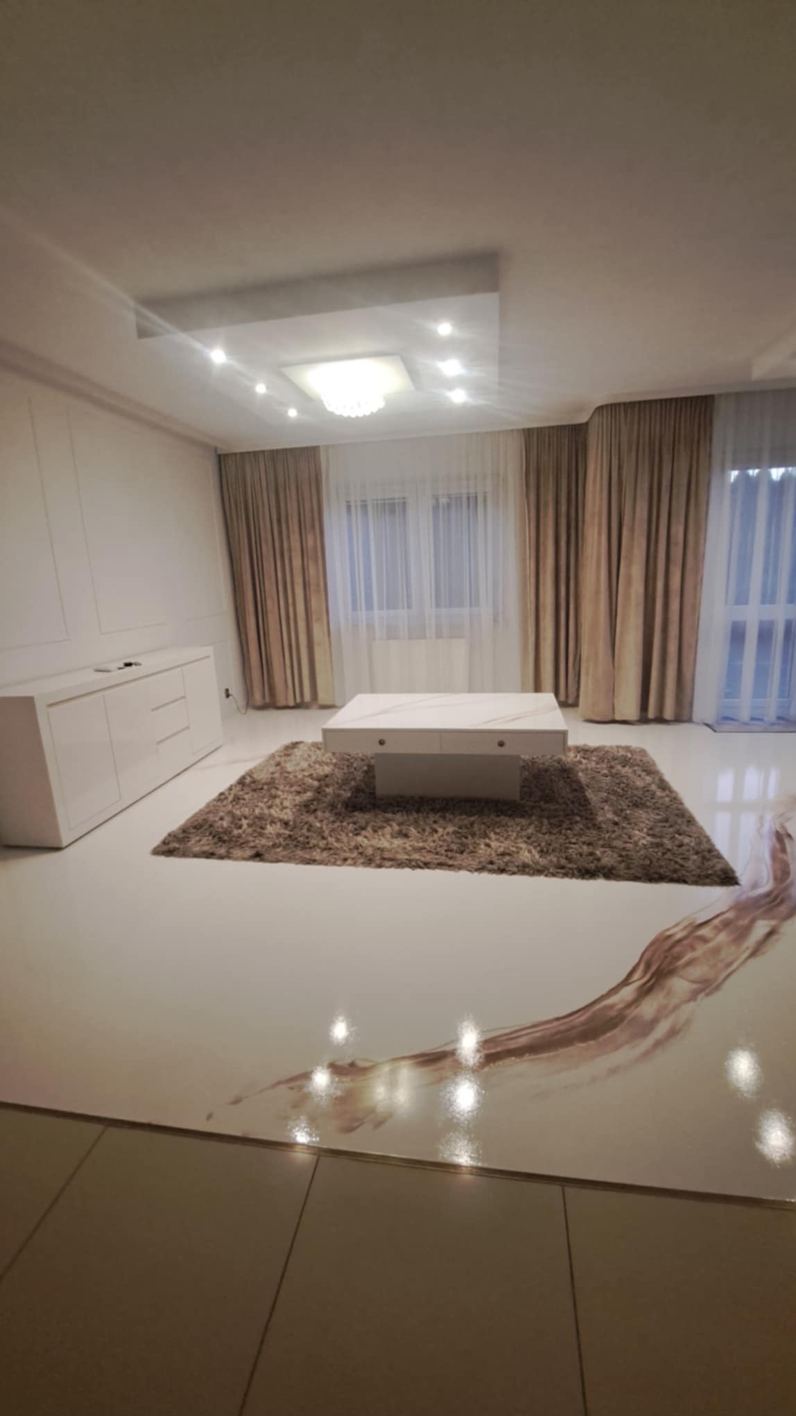 Jasne wnętrze salonu z żywiczną posadzką imitującą wzór płynącej wody, kremowe zasłony, biała komoda i stolik kawowy na puszystym dywanie.