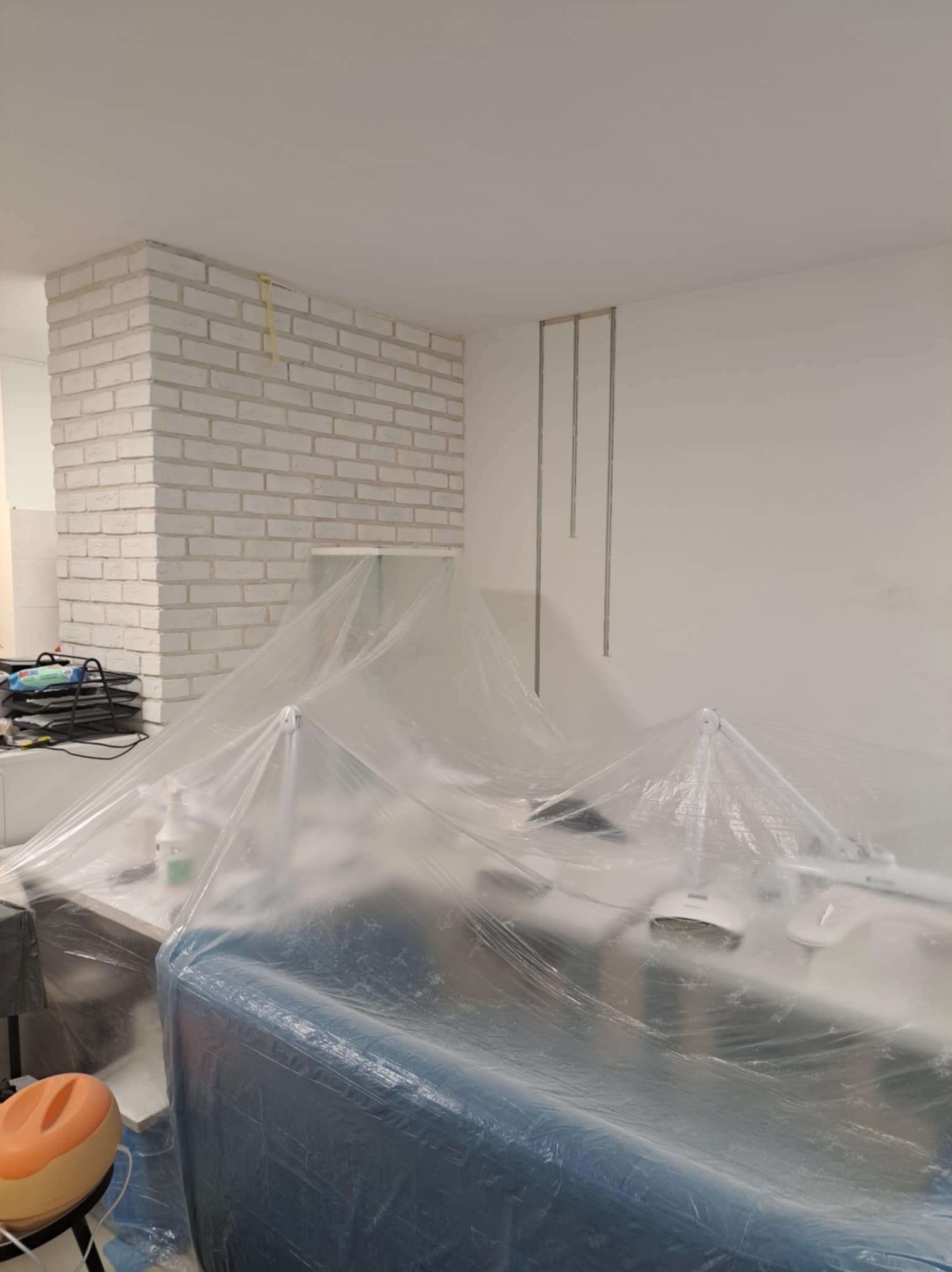Pomieszczenie przygotowane do malowania, meble i urządzenia zabezpieczone folią, widoczny fragment ściany z dekoracyjną cegłą pomalowaną na biało oraz metalowe profile na ścianie.