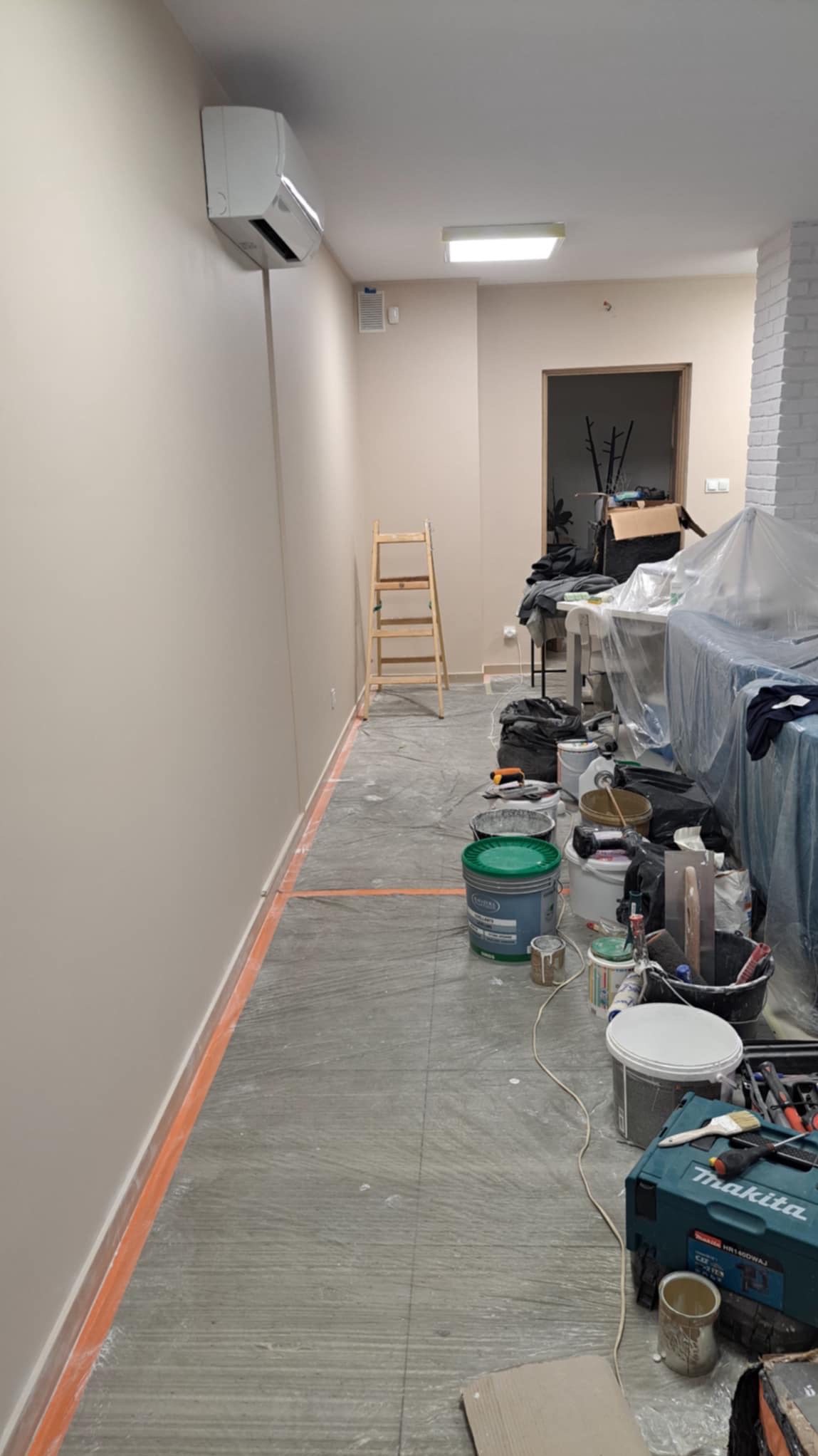 Pomieszczenie po malowaniu ścian na beżowo, widoczne ślady farby na podłodze zabezpieczonej folią, drabina, puszki z farbą i narzędzia malarskie.