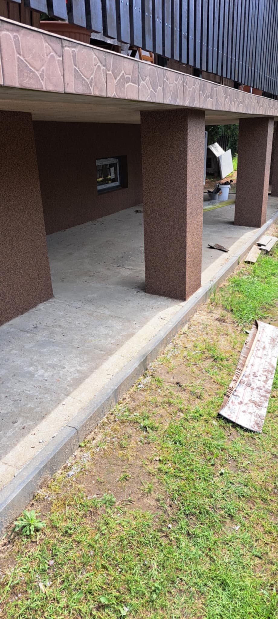 Wykończony podcień domu z brązową elewacją typu baranek, betonową podłogą i widocznymi elementami ogrodu.