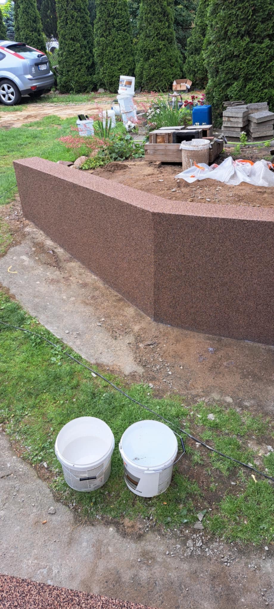 Wykończona, brązowa, ziarnista elewacja murku rabaty kwiatowej w ogrodzie, widoczne dwa białe wiadra z farbą i elementy roślinności w tle.