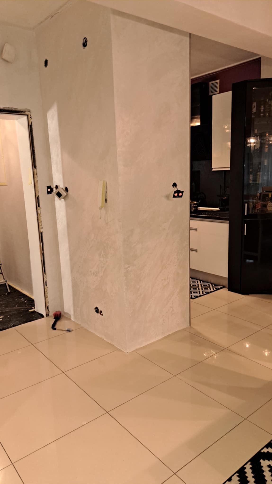 Ściana po nałożeniu gładzi dekoracyjnej w odcieniach szarości, widoczne puszki elektryczne, narzędzia malarskie i fragment wykończonej podłogi z płytek ceramicznych