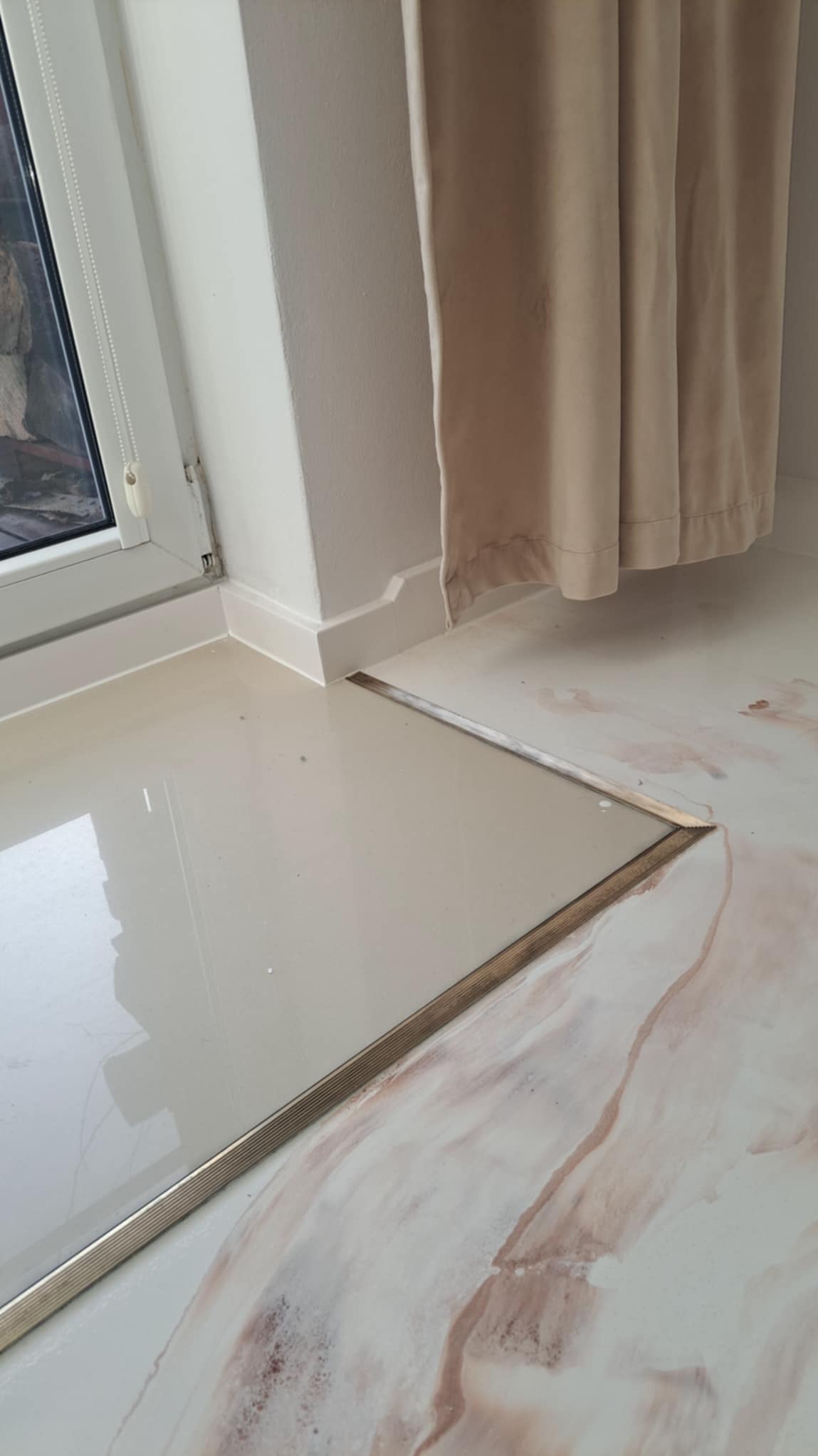 Połączenie dwóch rodzajów podłóg: jasnej, błyszczącej płytki z metalową listwą oraz podłogi żywicznej z marmurkowym wzorem w odcieniach bieli i brązu, widoczne przy oknie i zasłonie.