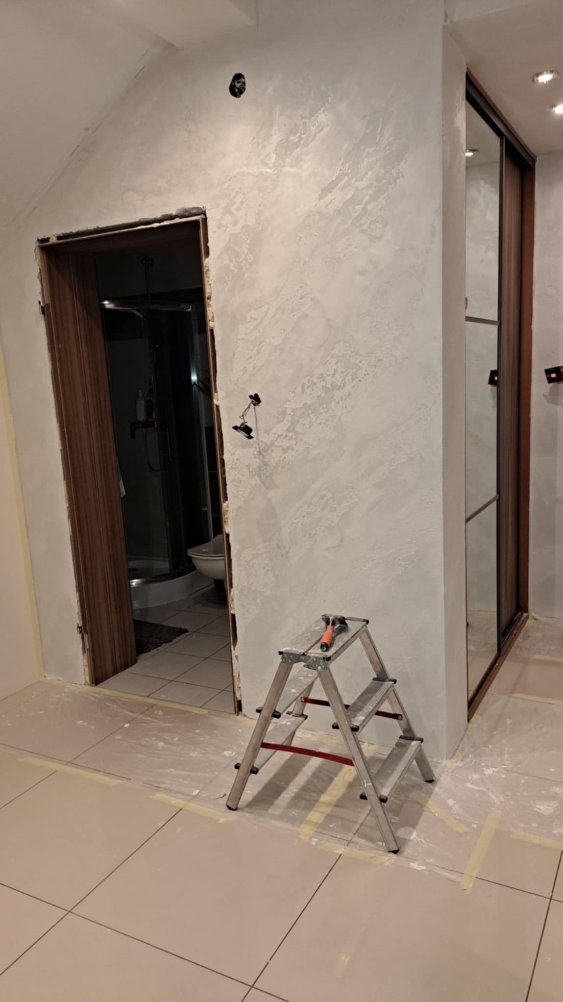 Ściana z dekoracyjną, jasnoszarą gładzią strukturalną w trakcie prac, widoczna drabina aluminiowa, zabezpieczona podłoga, otwarte drzwi do łazienki i fragment szafy przesuwnej.