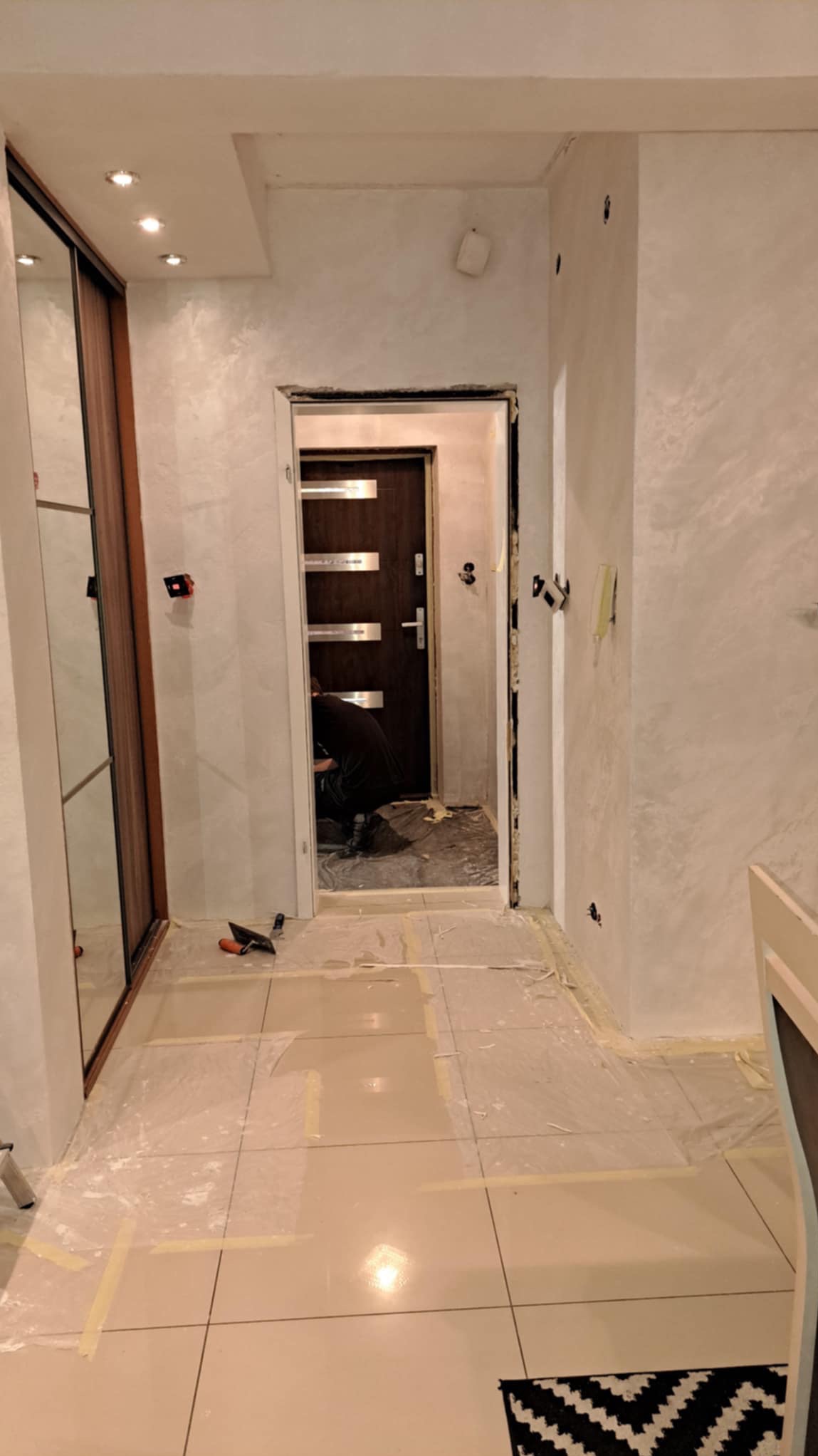 Korytarz w trakcie prac: ściany pokryte dekoracyjną gładzią w odcieniach bieli i szarości, zabezpieczona podłoga z jasnych płytek, widoczne narzędzia i sylwetka osoby pracującej przy drzwiach...