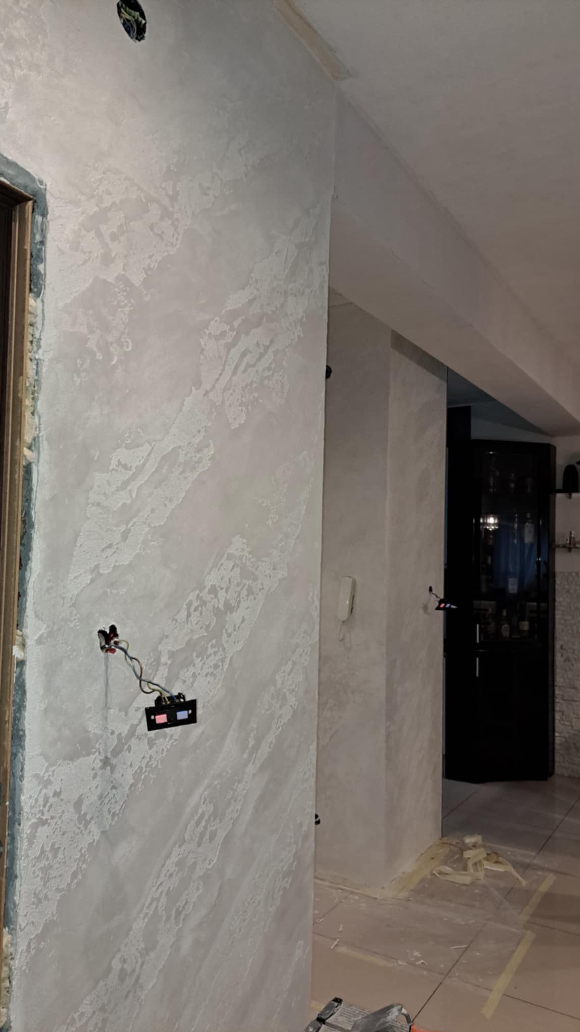 Ściana pokryta dekoracyjną, jasną gładzią w stylu weneckim, widoczne niedokończone wykończenia przy framudze drzwi i wystające przewody elektryczne.