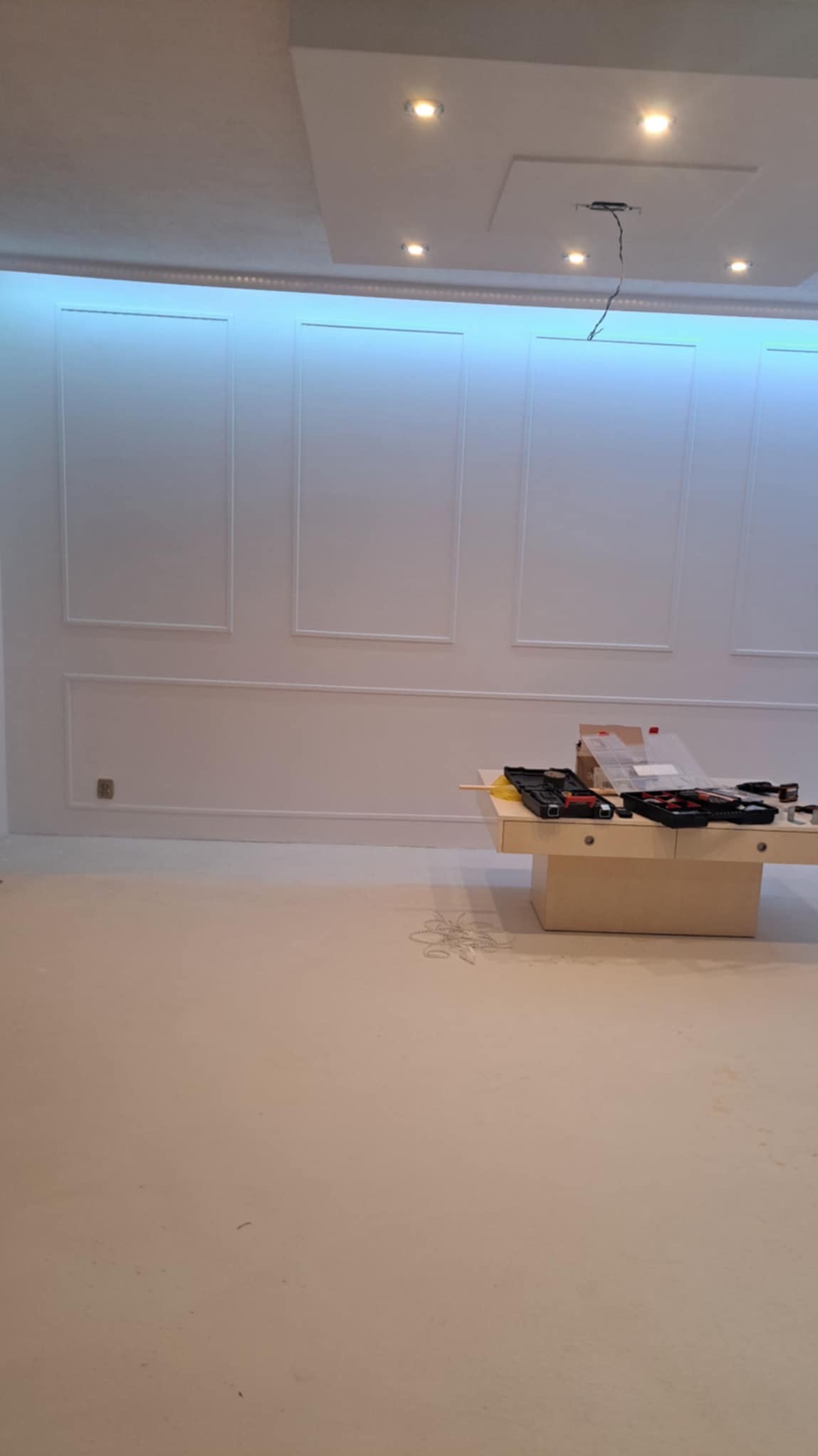 Białe ściany z dekoracyjnymi listwami i oświetleniem LED na suficie, narzędzia na stole w trakcie prac wykończeniowych.