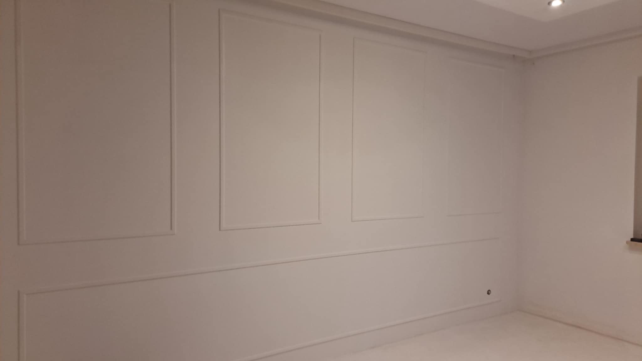 Pomalowane na biało ściany z dekoracyjnymi listwami w pustym pokoju, widoczny fragment sufitu z oświetleniem punktowym.