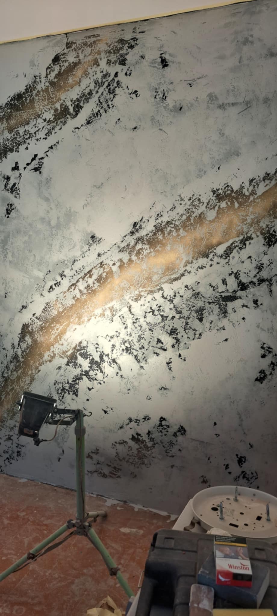 Ściana z dekoracyjną, szarą gładzią z nieregularnymi czarnymi plamami i złotym pasem, oświetlona lampą budowlaną na trójnogu, widoczny fragment podłogi i akcesoria malarskie.