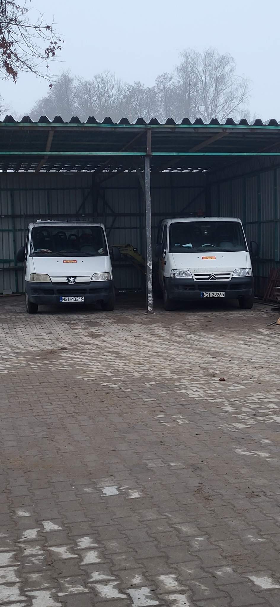 Dwa busy firmowe Peugeot i Citroen zaparkowane pod zadaszeniem, w tle widoczny mały sprzęt budowlany, prawdopodobnie minikoparka.