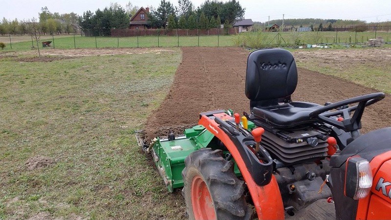 Traktor z glebogryzarką przygotowuje ziemię pod uprawę na polu w wiejskim otoczeniu, widoczny dom w tle.