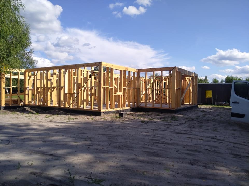 Wznoszenie szkieletu domu z drewna na fundamencie, widoczne czarne zabezpieczenie przeciwwilgociowe, w tle błękitne niebo z chmurami i fragment białego samochodu dostawczego.