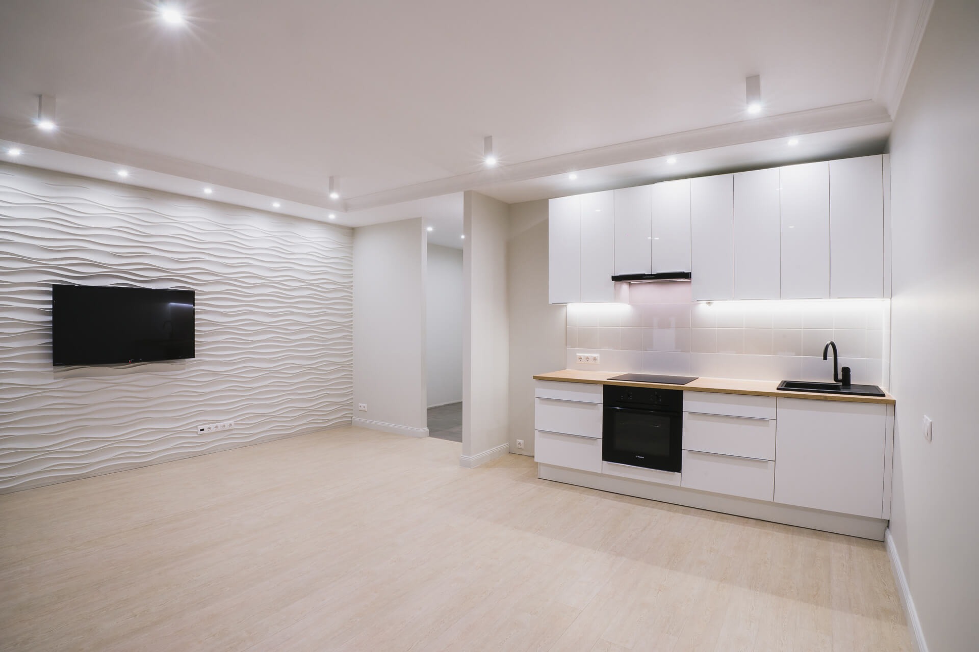 Nowoczesne wnętrze: białe, minimalistyczne meble kuchenne z drewnianym blatem, czarny zlew i płyta indukcyjna, ściana z dekoracyjną fakturą 3D oraz telewizor, jasna podłoga.