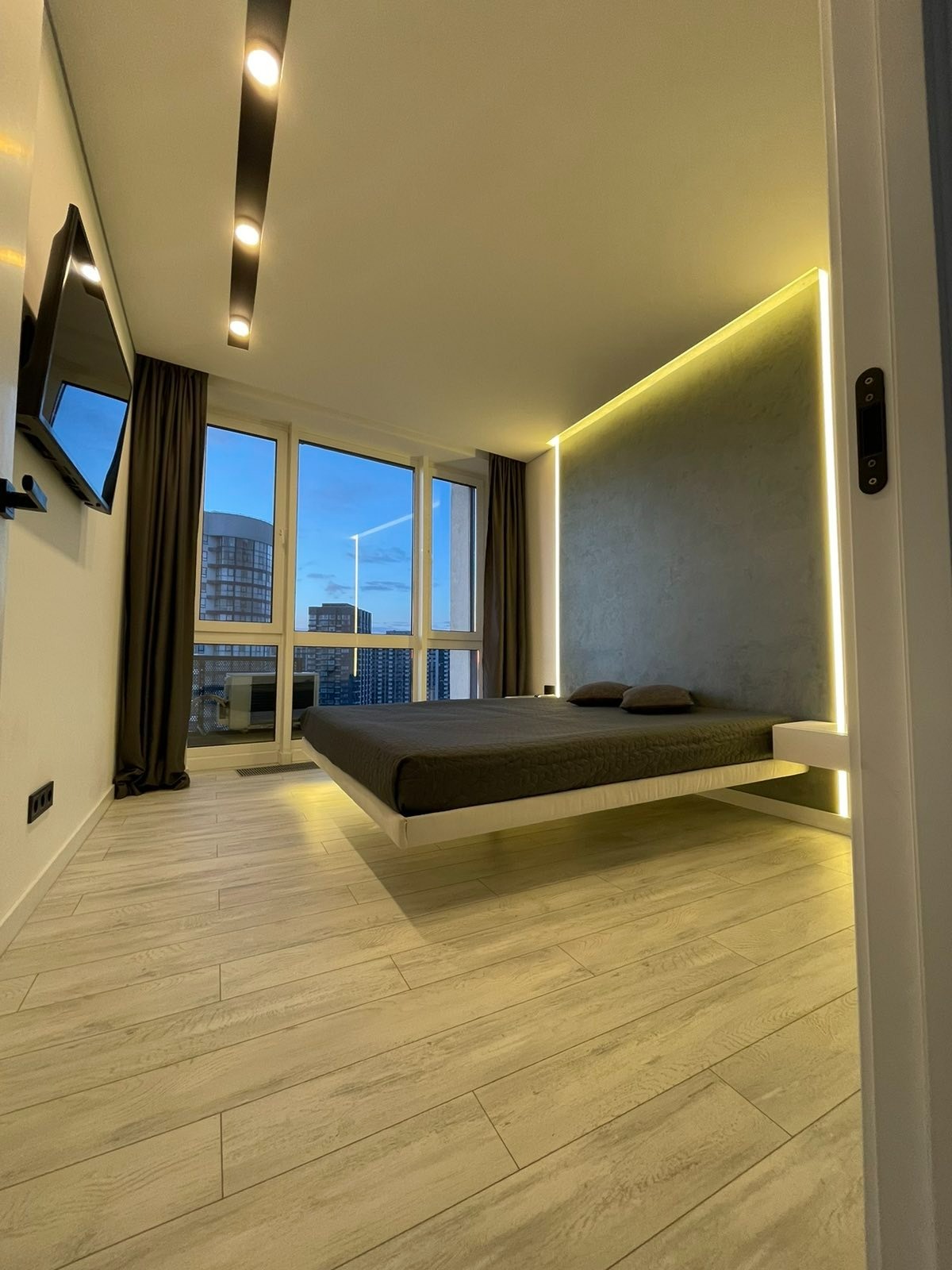 Nowoczesna sypialnia z podświetlanym łóżkiem, szarą ścianą z pionowym oświetleniem LED, parkietem i widokiem na miasto za oknem.