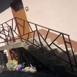 Uslugi spawalniczo monterskie - Betonowe schody zewnętrzne z geometryczną, brązową barierką. Pod schodami rower i torba. Dom z beżową elewacją i brązowym pasem. Błękitne niebo w tle.