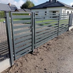 Uslugi spawalniczo monterskie - Nowoczesna, pozioma brama wjazdowa z metalowymi przęsłami w kolorze grafitowym, z widocznym zamkiem i fragmentem ogrodzenia panelowego. Tło stanowi dom jednorodzinny.