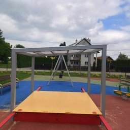 Uslugi spawalniczo monterskie - Metalowa konstrukcja z zadaszeniem nad piaskownicą i huśtawką na placu zabaw. Szara rama, żółta piaskownica, czerwona podstawa. W tle dom i zieleń.