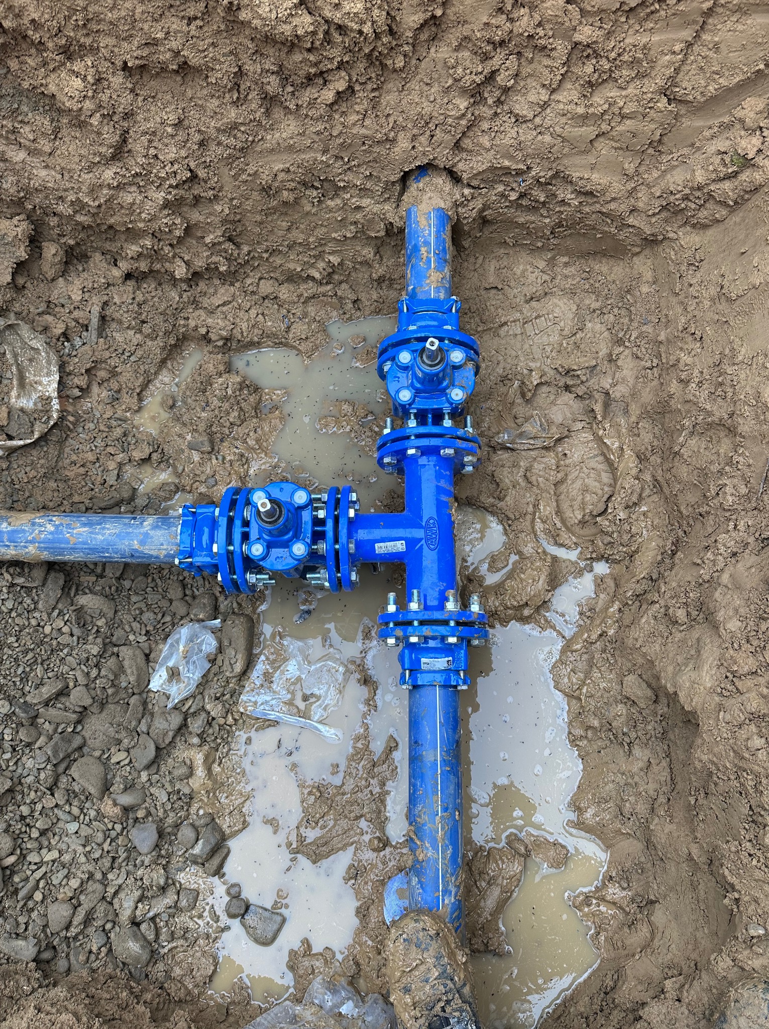 Niebieski, stalowy trójnik z zaworami w wykopie, otoczony błotem i drobnymi kamieniami. Widoczne ślady kopania. Instalacja wodociągowa w trakcie montażu.