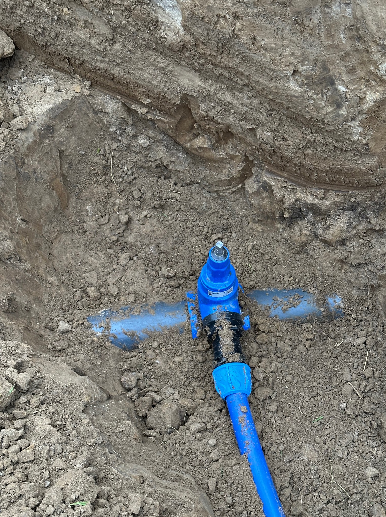 Niebieskie rury wodociągowe z zaworem w wykopie ziemnym, widoczne z góry. Instalacja przyłącza wodociągowego w trakcie realizacji.