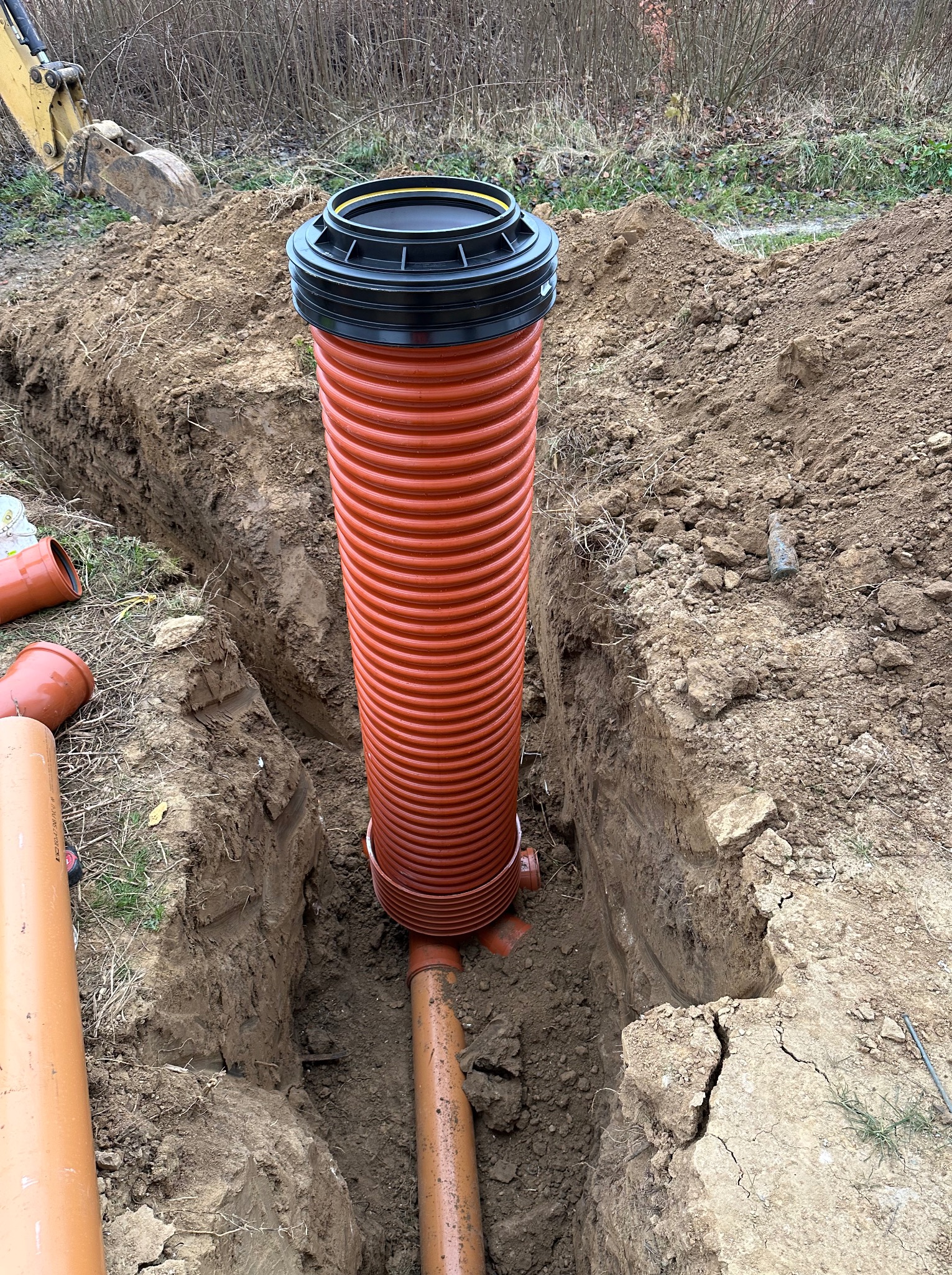 Montaż pomarańczowej rury kanalizacyjnej w wykopie ziemnym, z widoczną czarną pokrywą i fragmentem żółtej koparki w tle. Etap prac ziemnych i podłączenia.