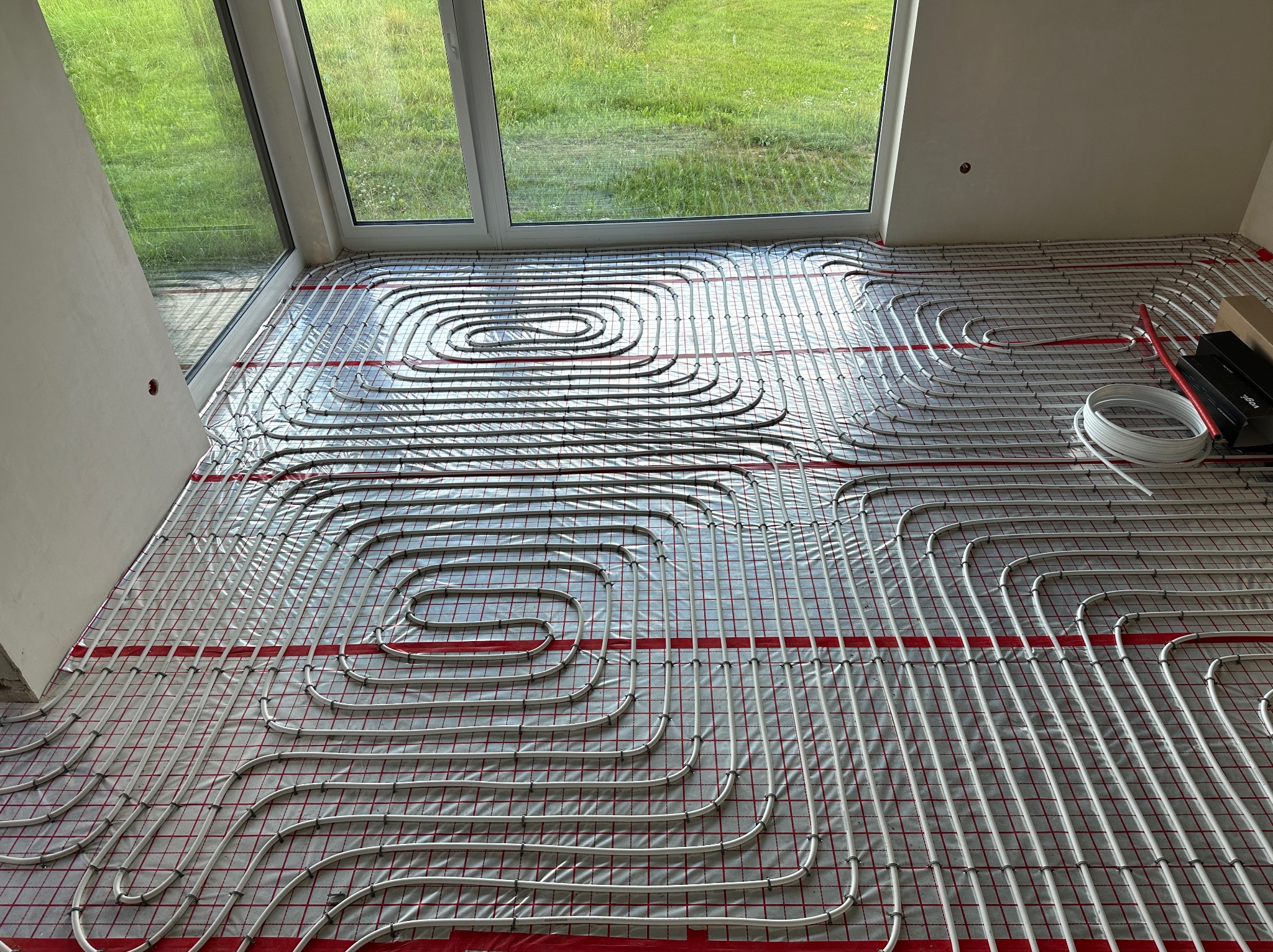 Białe rury ogrzewania podłogowego ułożone na czerwonej folii z siatką, widok z góry, z oknem w tle. Etap instalacji przed wylaniem posadzki.