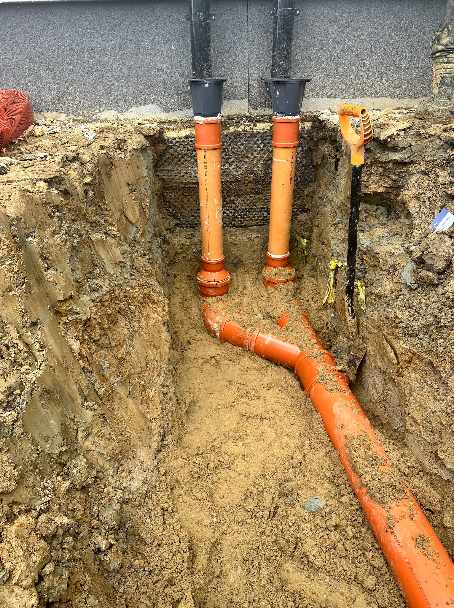 Pomarańczowe rury kanalizacyjne w wykopie, widoczne połączenia i fragment ściany budynku. Szpadel wbity w ziemię. Prace instalacyjne w toku.