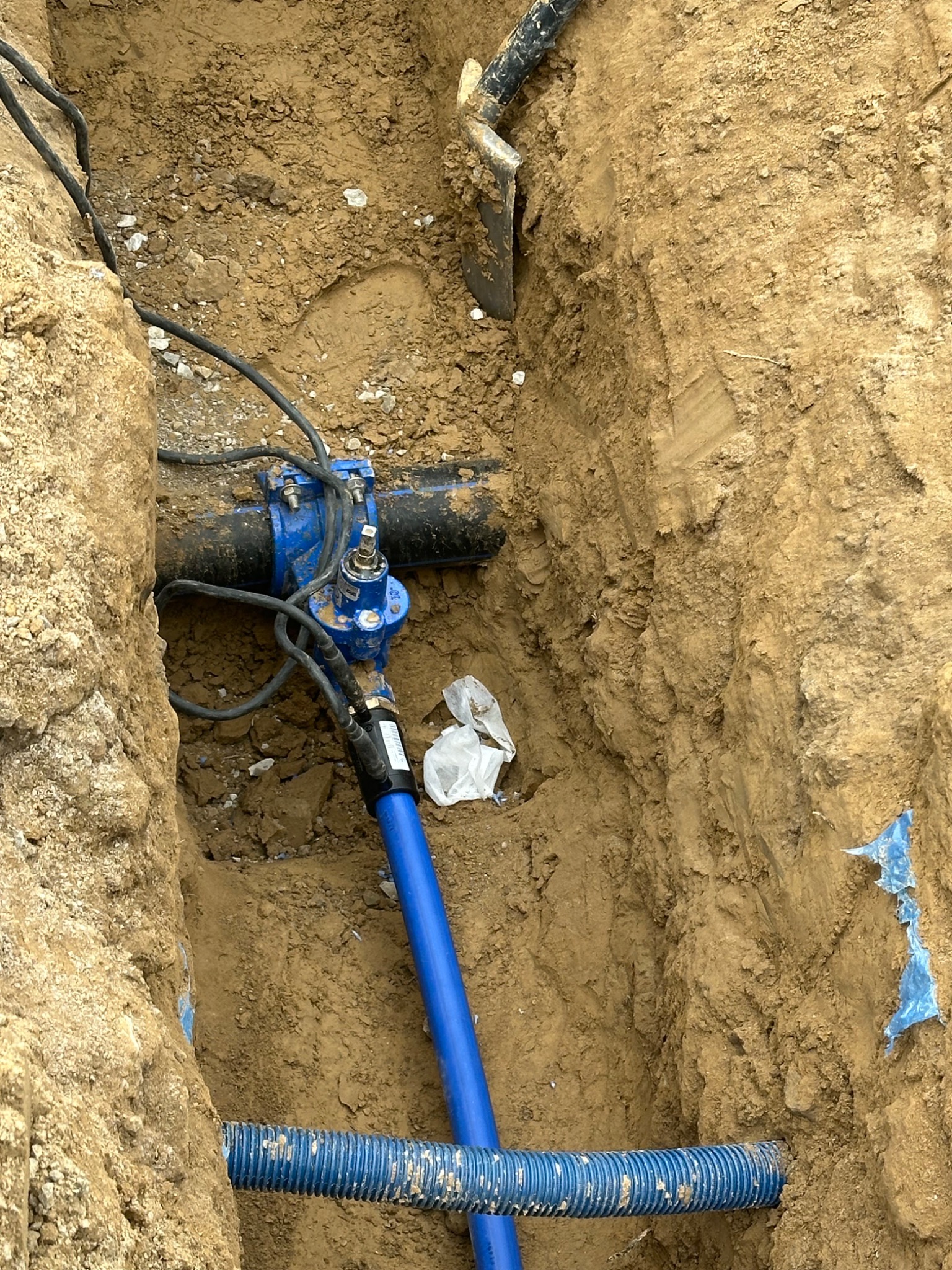 Niebieska rura wodociągowa w wykopie ziemnym, z widocznymi złączkami i czarnym kablem. Fragment łopaty w tle. Wykopane ściany z piasku.