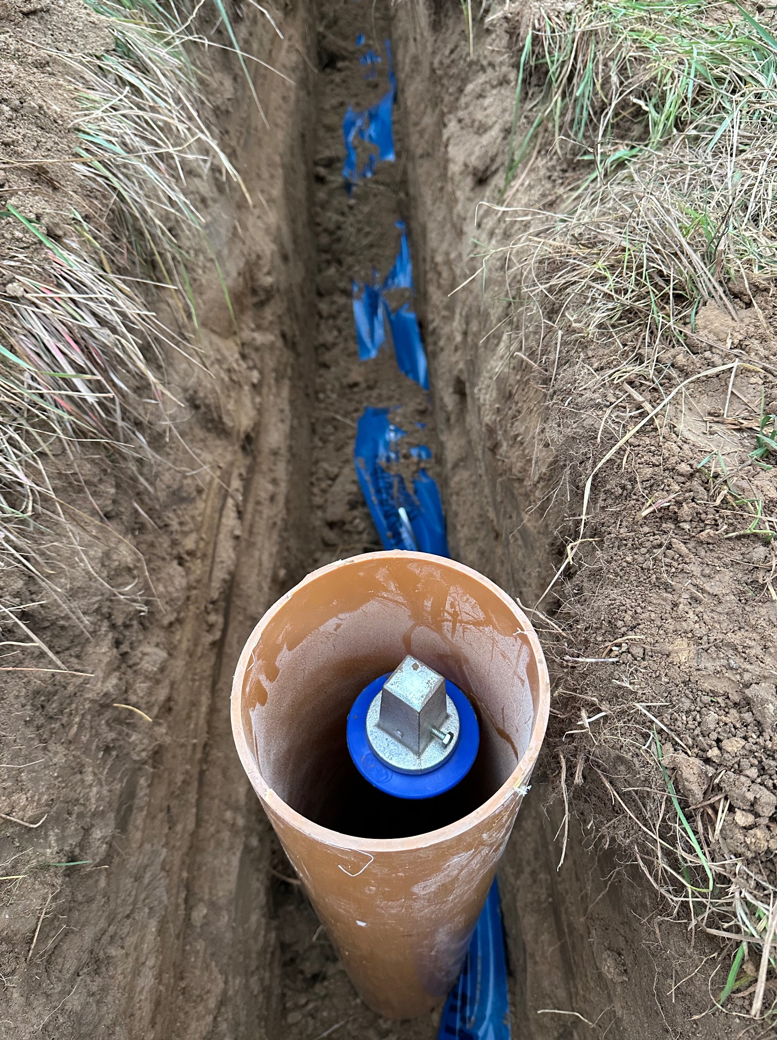 Brązowa rura pionowa w wykopie z niebieskimi rurami w ziemi. Widok z góry na instalację wodno-kanalizacyjną podczas prac ziemnych.