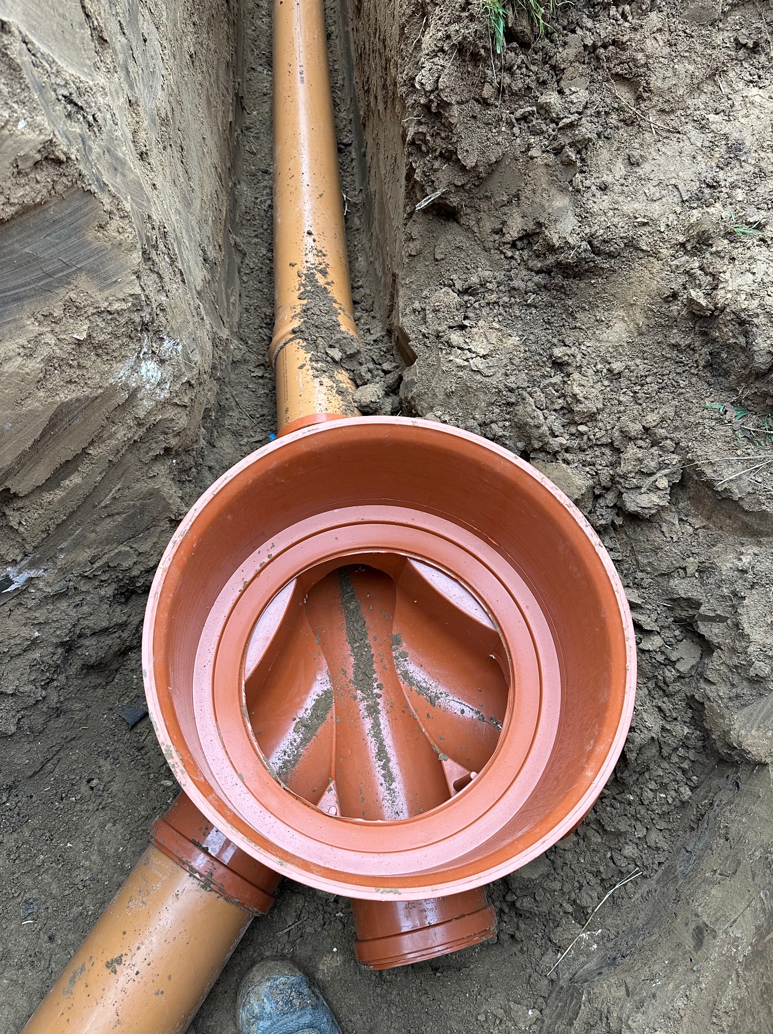 Brązowa studzienka kanalizacyjna z rurami w wykopie ziemnym, widok z góry, fragment buta roboczego w lewym dolnym rogu kadru.
