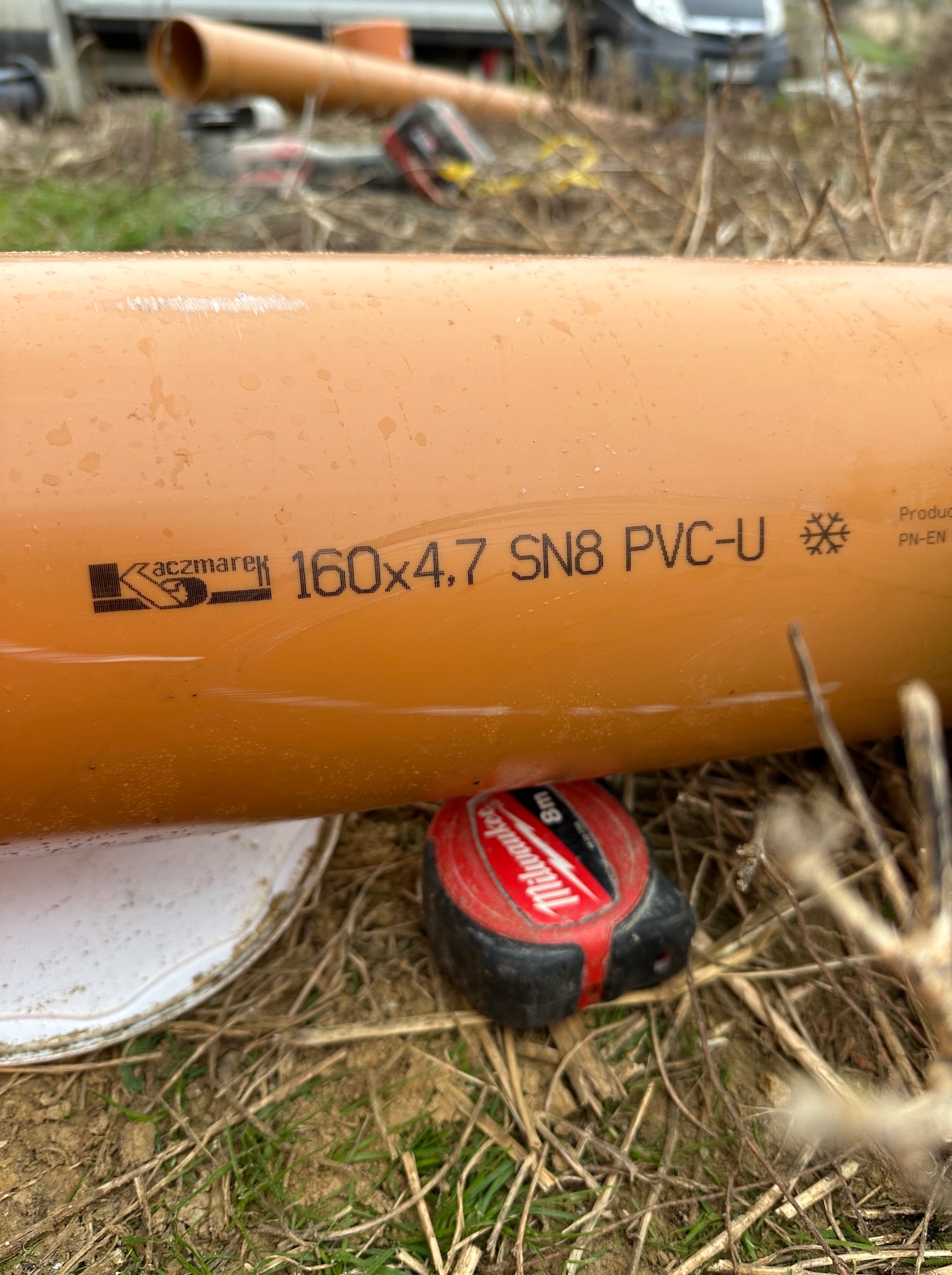 Pomarańczowa rura PVC-U Kaczmarek 160x4.7 SN8 leży na ziemi obok miarki Milwaukee 8m. W tle inne rury i pojazd. Widoczne oznaczenia producenta.