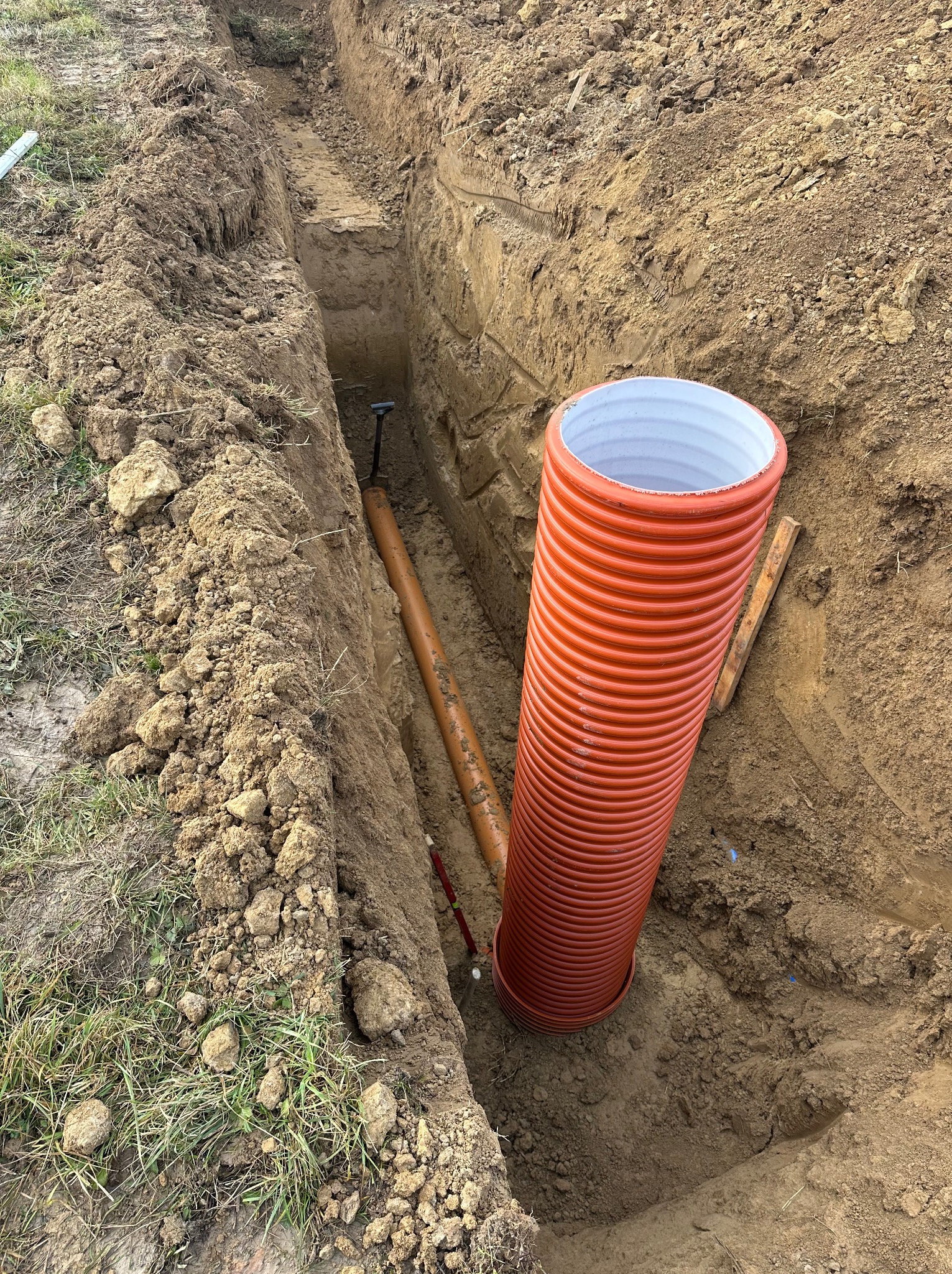 Wykop z pomarańczową rurą karbowaną i brązową rurą gładką w środku. Widoczny fragment łopaty. Grunt ziemny, gliniasty.