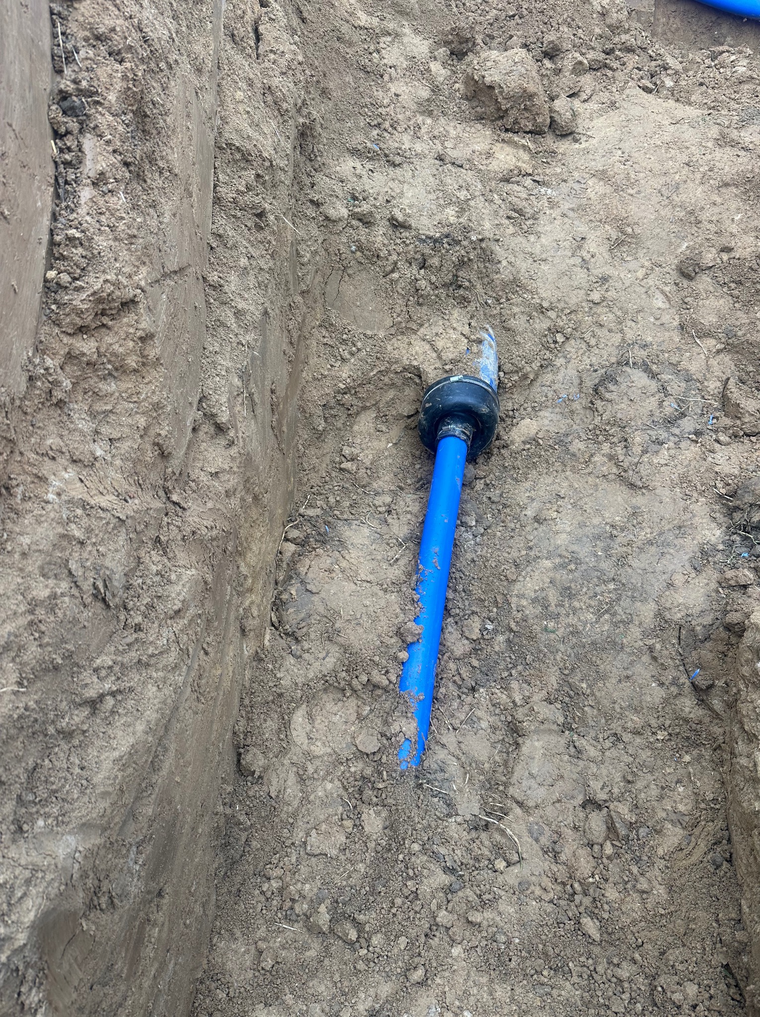 Niebieska rura wodociągowa z czarnym łącznikiem w wykopie ziemnym. Widok z góry na instalację wodną w trakcie prac ziemnych.