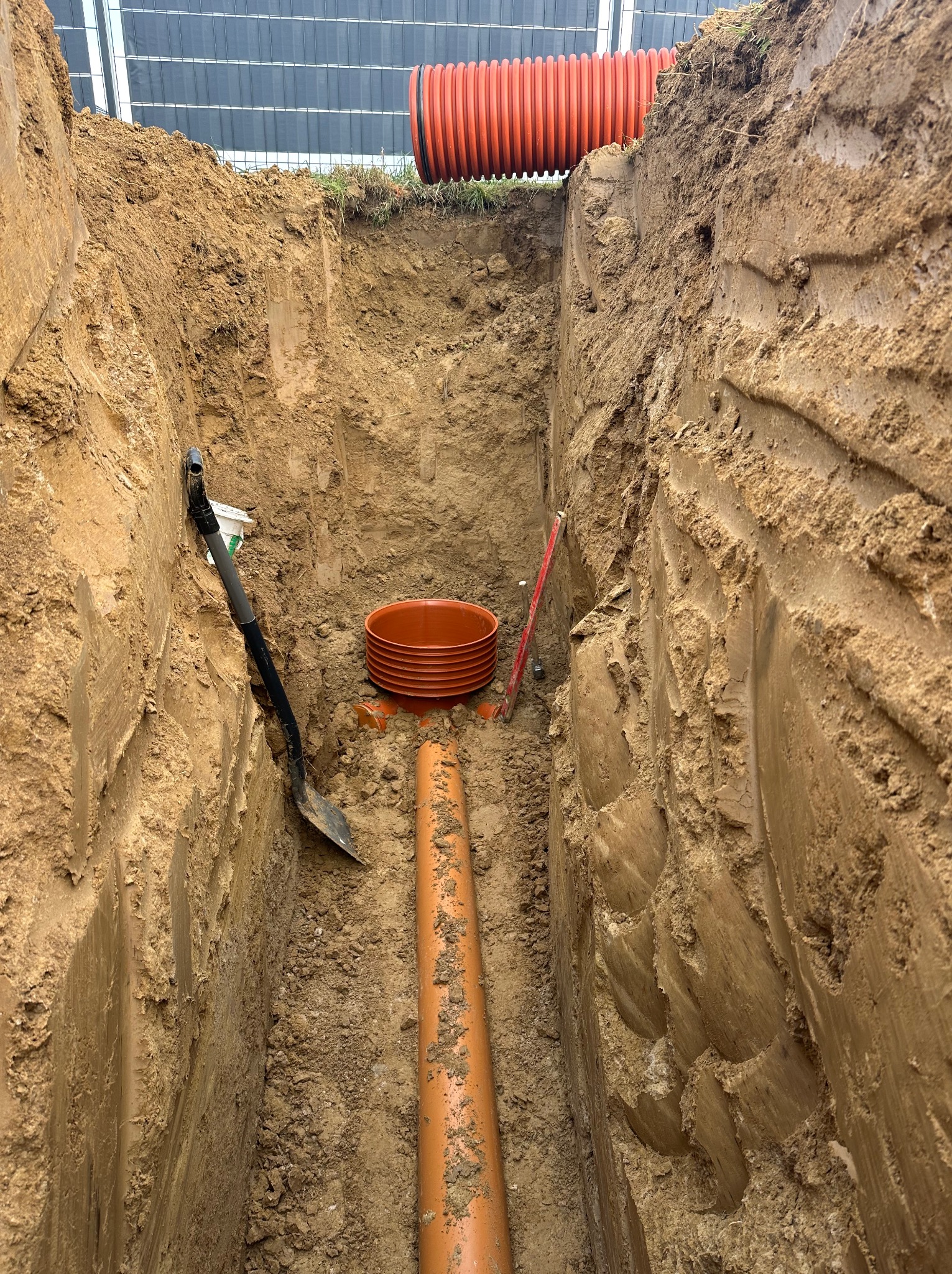 Wykop z pomarańczowymi rurami kanalizacyjnymi w ziemi, widoczna łopata i poziomica. Rury karbowane na powierzchni terenu. Budynek w tle.