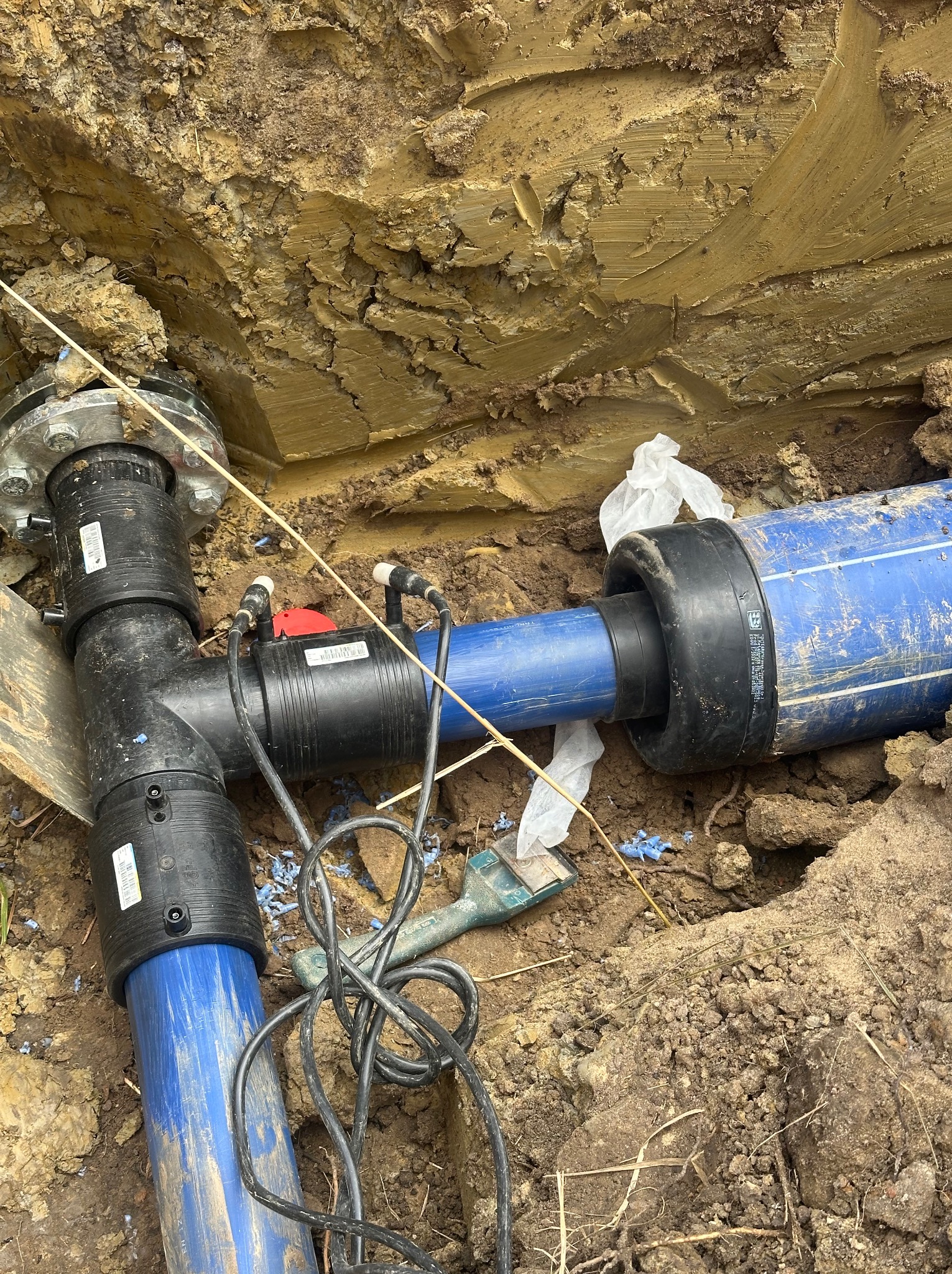 Niebieskie rury połączone czarnym trójnikiem z kołnierzem w wykopie ziemnym. Widoczne przewody i narzędzia. Instalacja wodociągowa w trakcie montażu.