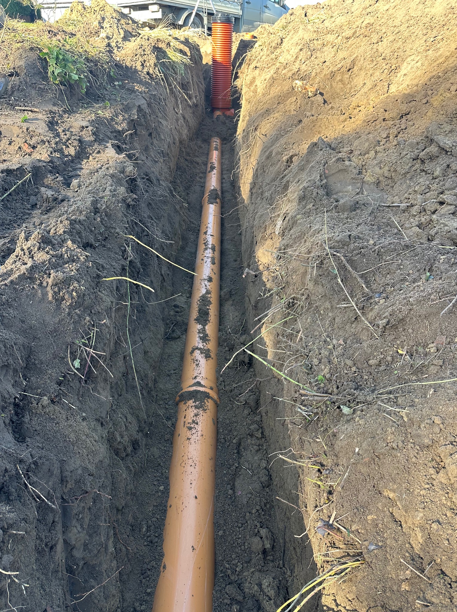 Brązowa rura kanalizacyjna ułożona w wykopie ziemnym, widok z góry. W tle widoczny fragment pomarańczowej rury pionowej i samochodu.