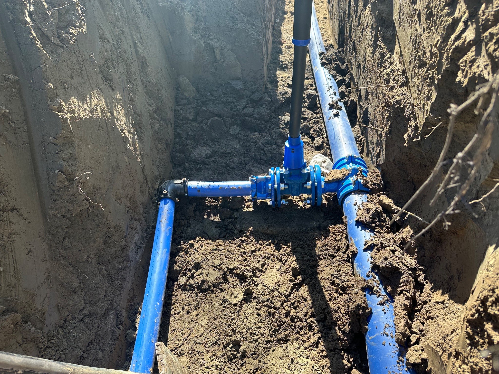Niebieskie rury wodociągowe w wykopie ziemnym, z widocznymi złączkami i zaworami. Instalacja w trakcie budowy, ziemia wokół rur.