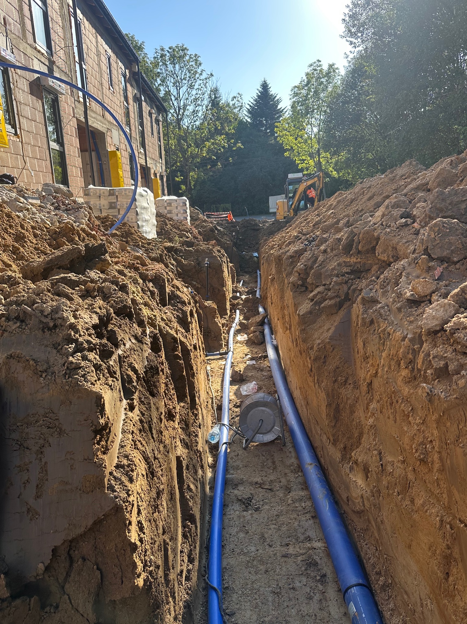 Wykop z ułożonymi niebieskimi rurami w Boguchwale, prawdopodobnie instalacja wodociągowa. W tle budynek w trakcie budowy i koparka.