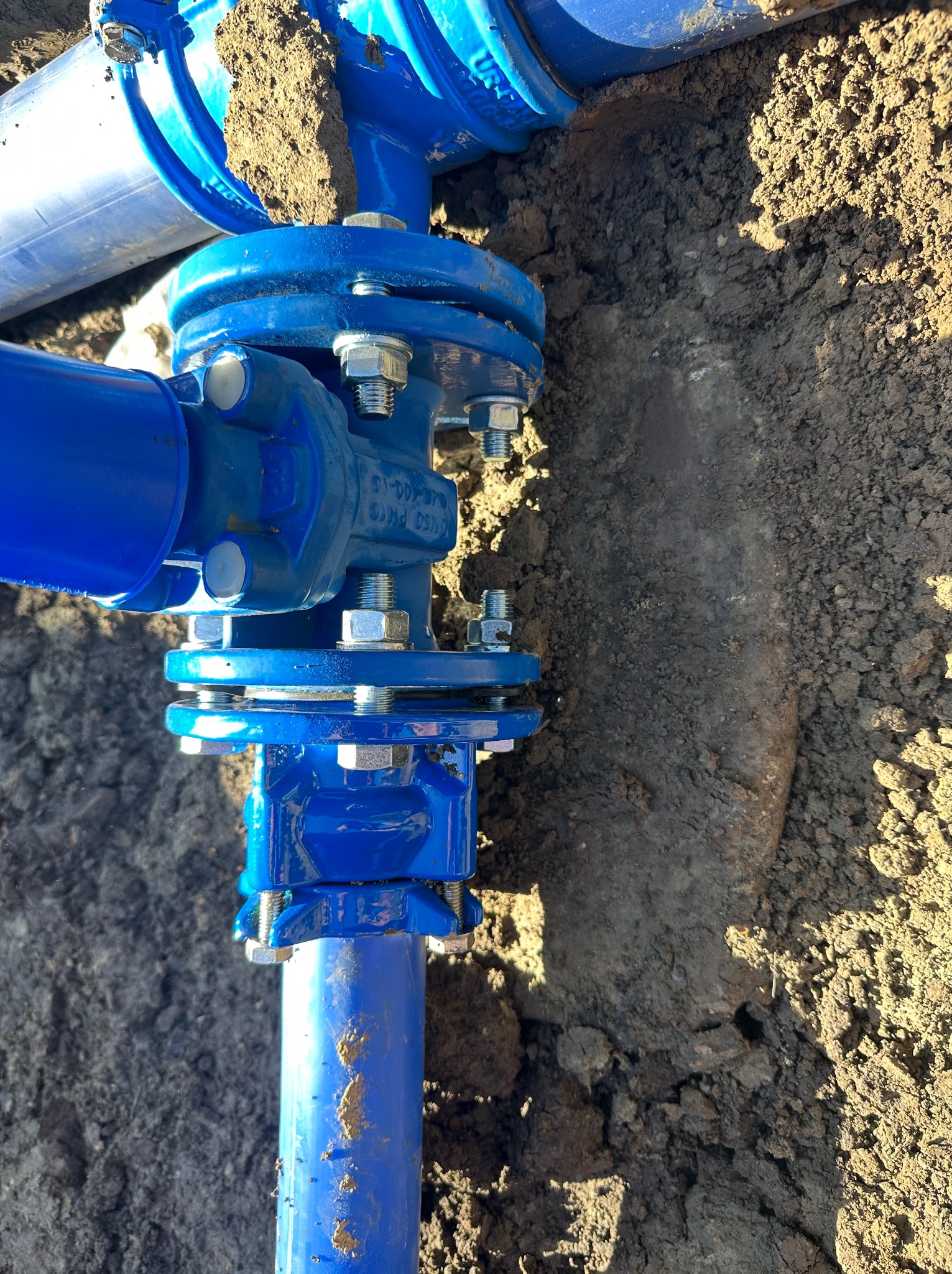 Niebieskie rury wodociągowe z łącznikami śrubowymi, częściowo zakopane w ziemi. Widoczne ślady świeżych wykopów i ziemi na elementach instalacji.