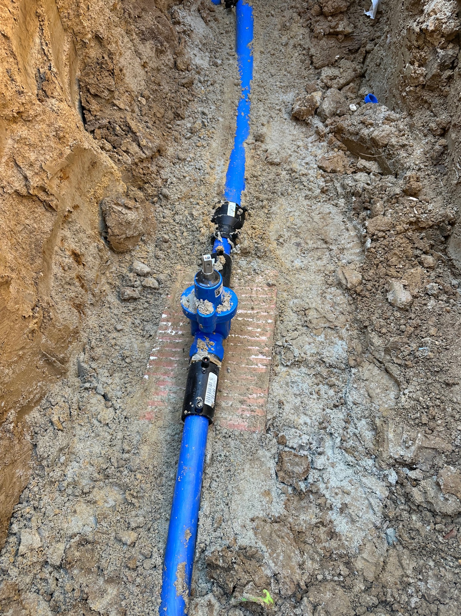 Niebieska rura wodociągowa z zaworem w wykopie ziemnym, widok z góry. Fragment muru z cegieł pod zaworem. Instalacja przyłącza wodociągowego w trakcie prac.