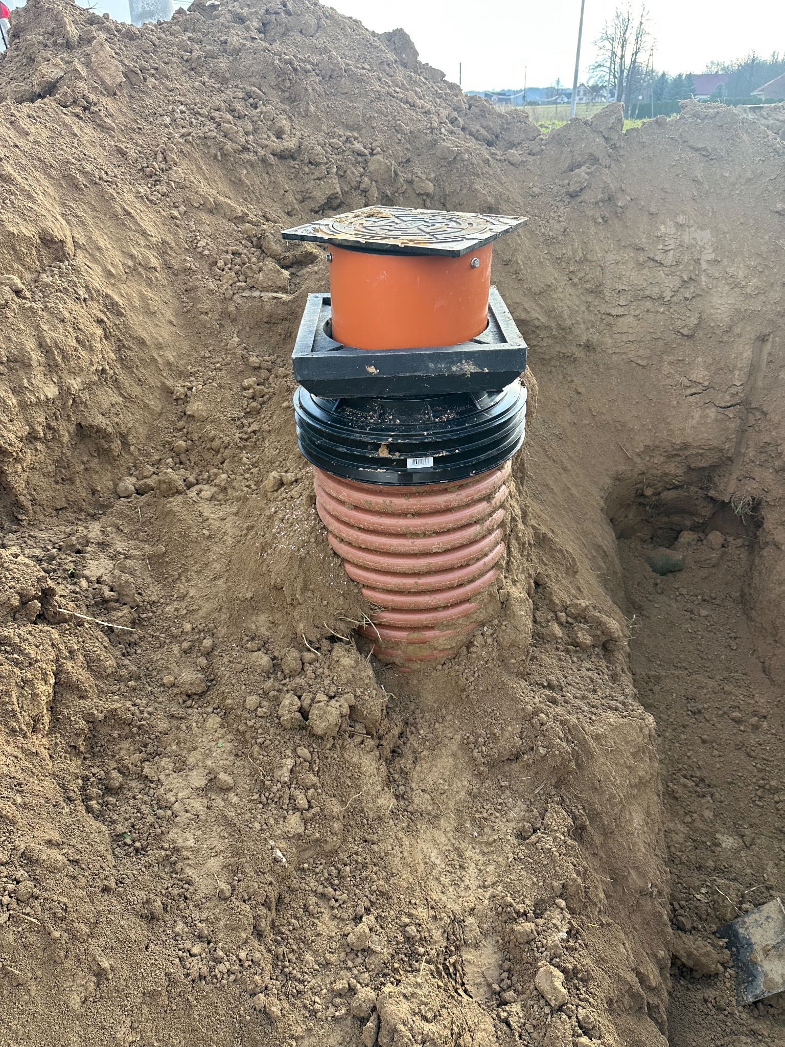 Montaż pomarańczowej studzienki kanalizacyjnej z czarnym włazem w wykopie ziemnym. Widoczne warstwy gruntu i fragment łopaty. Prace ziemne w toku.