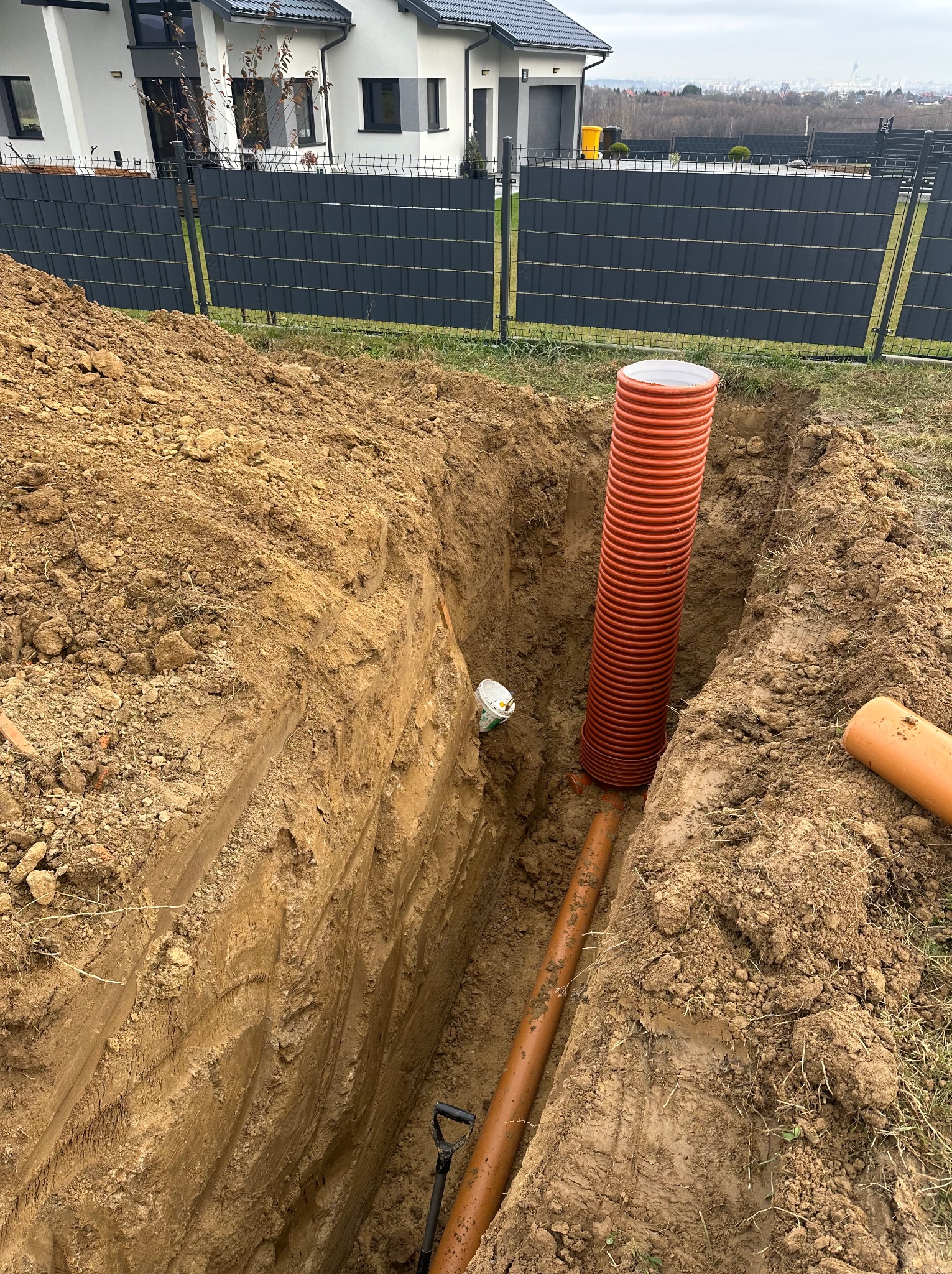 Wykop z pomarańczową rurą karbowaną pionowo i rurą gładką poziomo. Widoczny fragment szpadla, ziemia i ogrodzenie posesji w tle.