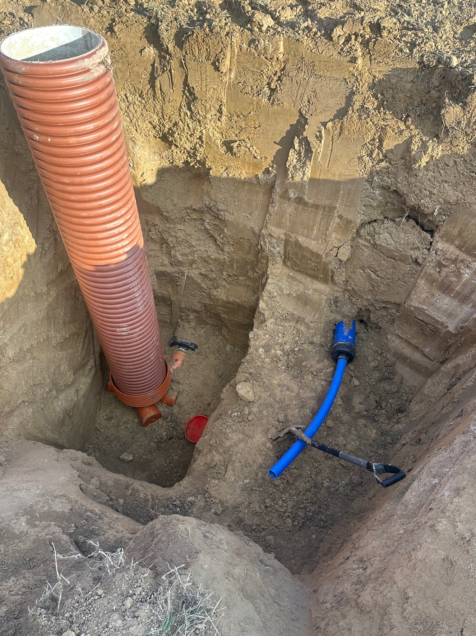 Wykop z pomarańczową rurą karbowaną i niebieską rurą PE, widoczne narzędzia. Instalacja w ziemi, etap prac ziemnych.