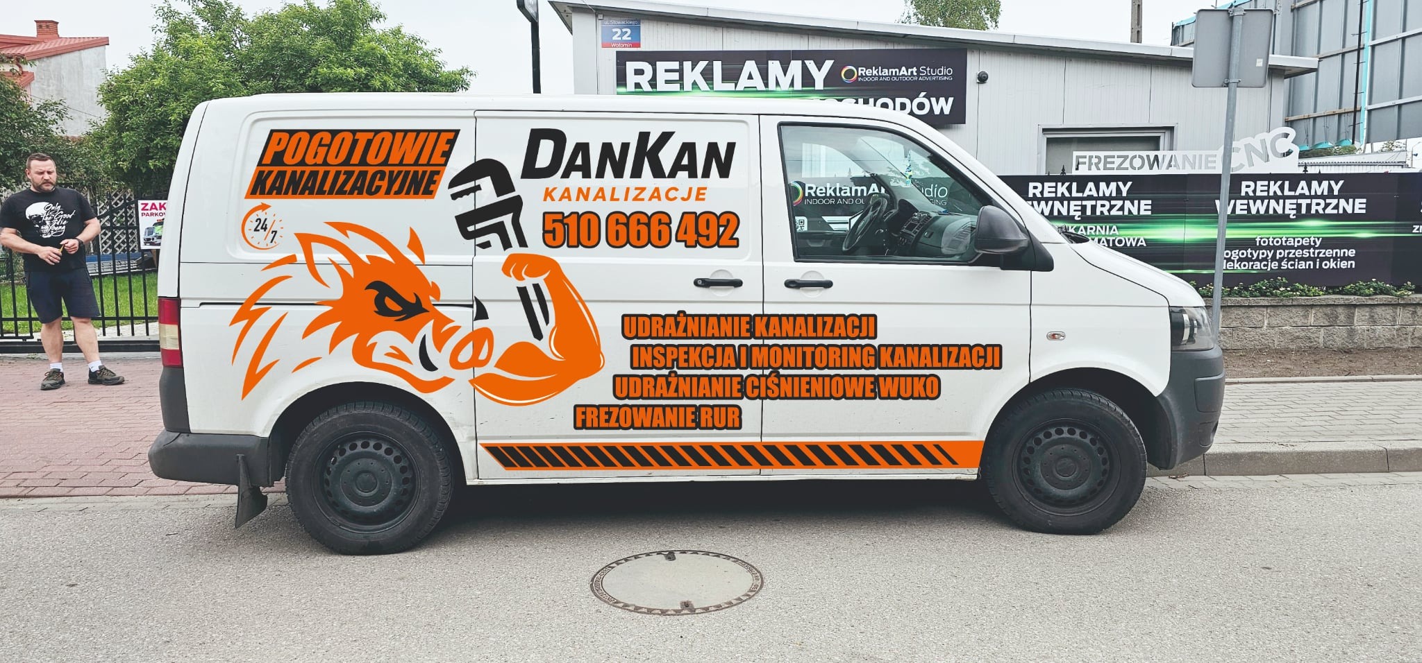 Biały furgon z grafiką pomarańczowego wilka i napisem 'DanKan Kanalizacje 510 666 492' na tle ulicy i budynku z reklamami.
