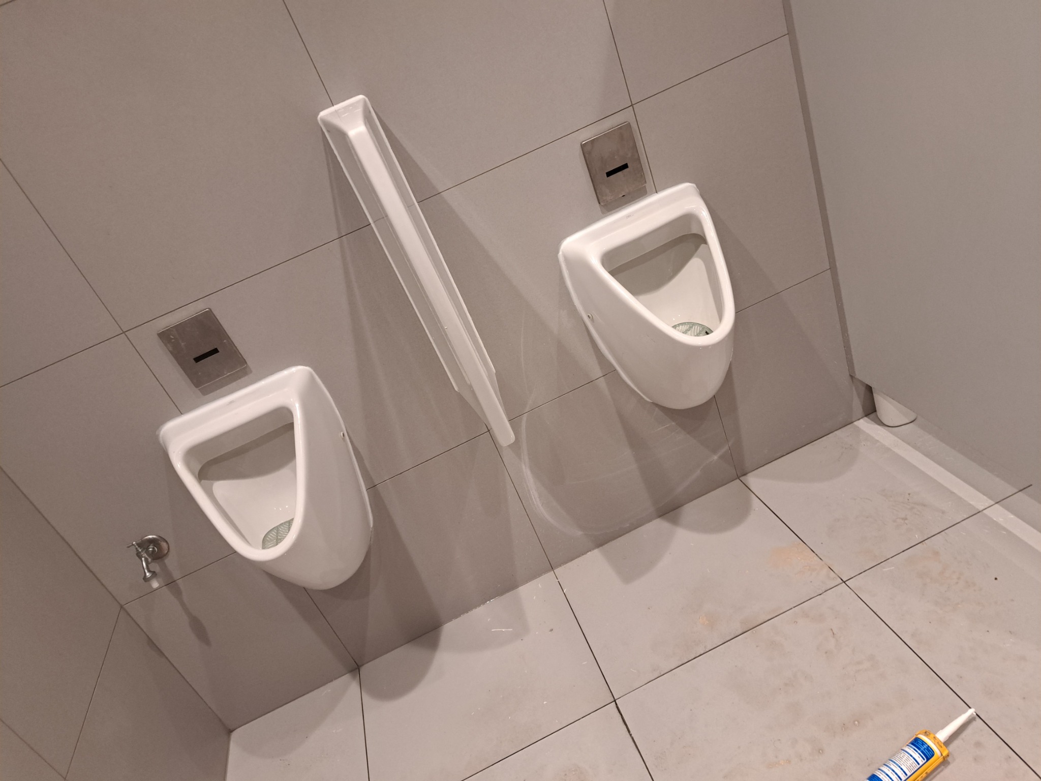 Dwa białe pisuary na szarej ścianie z płytkami, jeden z zamontowanym odpływem. Na podłodze leży tuba silikonu, co sugeruje prace hydrauliczne.