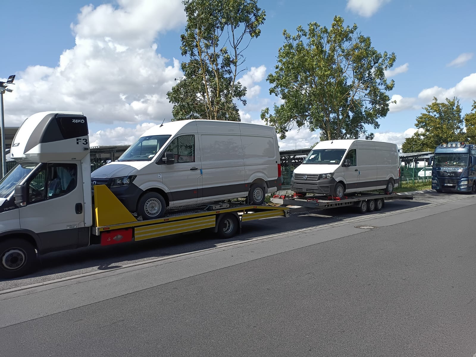 Laweta przewozi dwa białe busy na żółtej platformie. W tle drzewa i niebieski samochód ciężarowy. Dzień, pochmurne niebo.